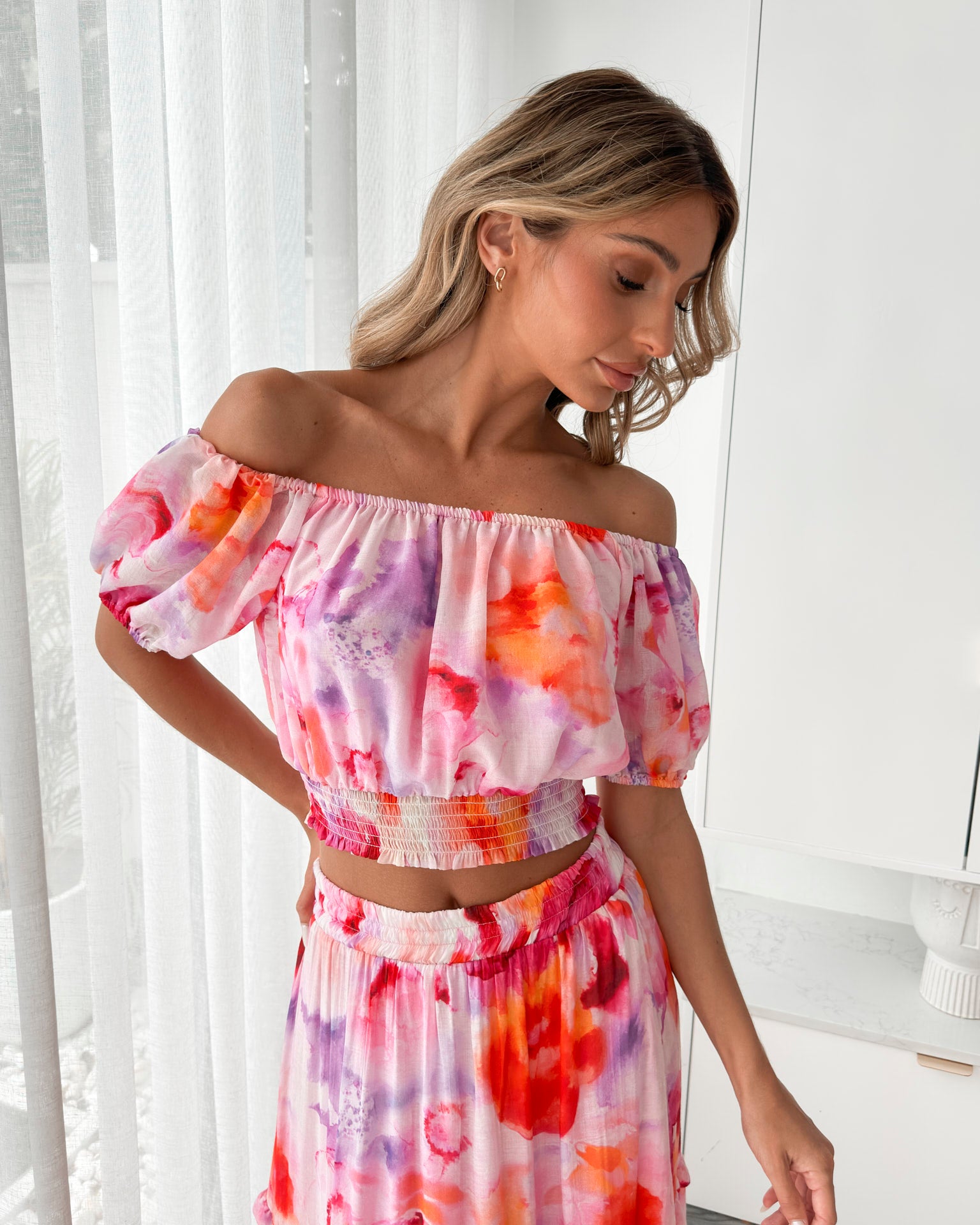 A woman stands indoors in a well-lit, modern room with sheer white curtains and minimalist white cabinetry. She is wearing a colorful off-the-shoulder two-piece outfit consisting of a ruffled crop top and a flowing maxi skirt with a vibrant watercolor floral print in pink, purple, orange, and red hues. Her wavy hair is styled loosely, and she stands gracefully with her hands gently clasped in front, exuding a fresh, summery elegance.