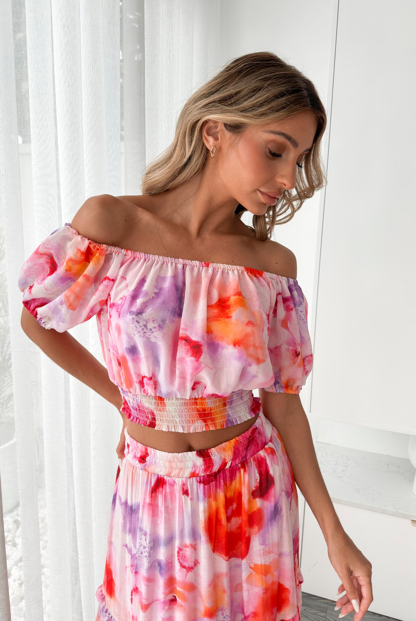 A woman stands indoors in a well-lit, modern room with sheer white curtains and minimalist white cabinetry. She is wearing a colorful off-the-shoulder two-piece outfit consisting of a ruffled crop top and a flowing maxi skirt with a vibrant watercolor floral print in pink, purple, orange, and red hues. Her wavy hair is styled loosely, and she stands gracefully with her hands gently clasped in front, exuding a fresh, summery elegance.