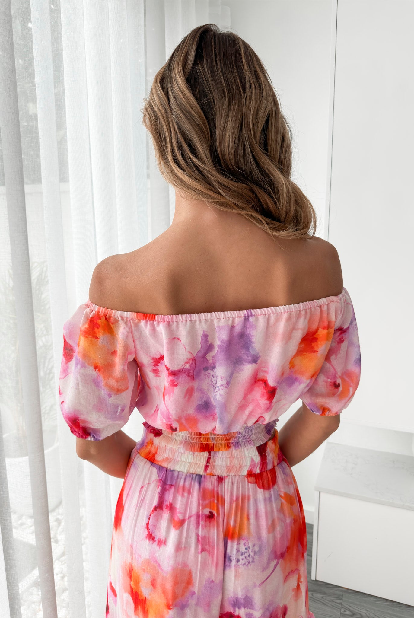 A woman stands indoors in a well-lit, modern room with sheer white curtains and minimalist white cabinetry. She is wearing a colorful off-the-shoulder two-piece outfit consisting of a ruffled crop top and a flowing maxi skirt with a vibrant watercolor floral print in pink, purple, orange, and red hues. Her wavy hair is styled loosely, and she stands gracefully with her hands gently clasped in front, exuding a fresh, summery elegance.