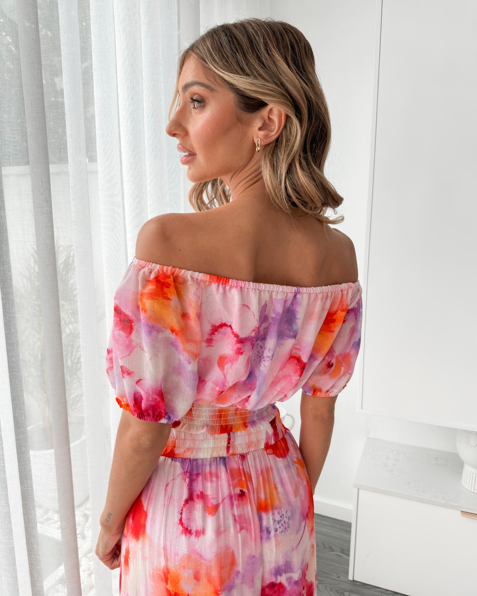 A woman stands indoors in a well-lit, modern room with sheer white curtains and minimalist white cabinetry. She is wearing a colorful off-the-shoulder two-piece outfit consisting of a ruffled crop top and a flowing maxi skirt with a vibrant watercolor floral print in pink, purple, orange, and red hues. Her wavy hair is styled loosely, and she stands gracefully with her hands gently clasped in front, exuding a fresh, summery elegance.