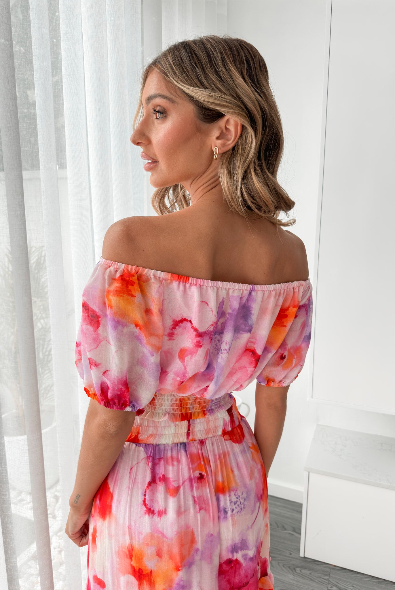 A woman stands indoors in a well-lit, modern room with sheer white curtains and minimalist white cabinetry. She is wearing a colorful off-the-shoulder two-piece outfit consisting of a ruffled crop top and a flowing maxi skirt with a vibrant watercolor floral print in pink, purple, orange, and red hues. Her wavy hair is styled loosely, and she stands gracefully with her hands gently clasped in front, exuding a fresh, summery elegance.