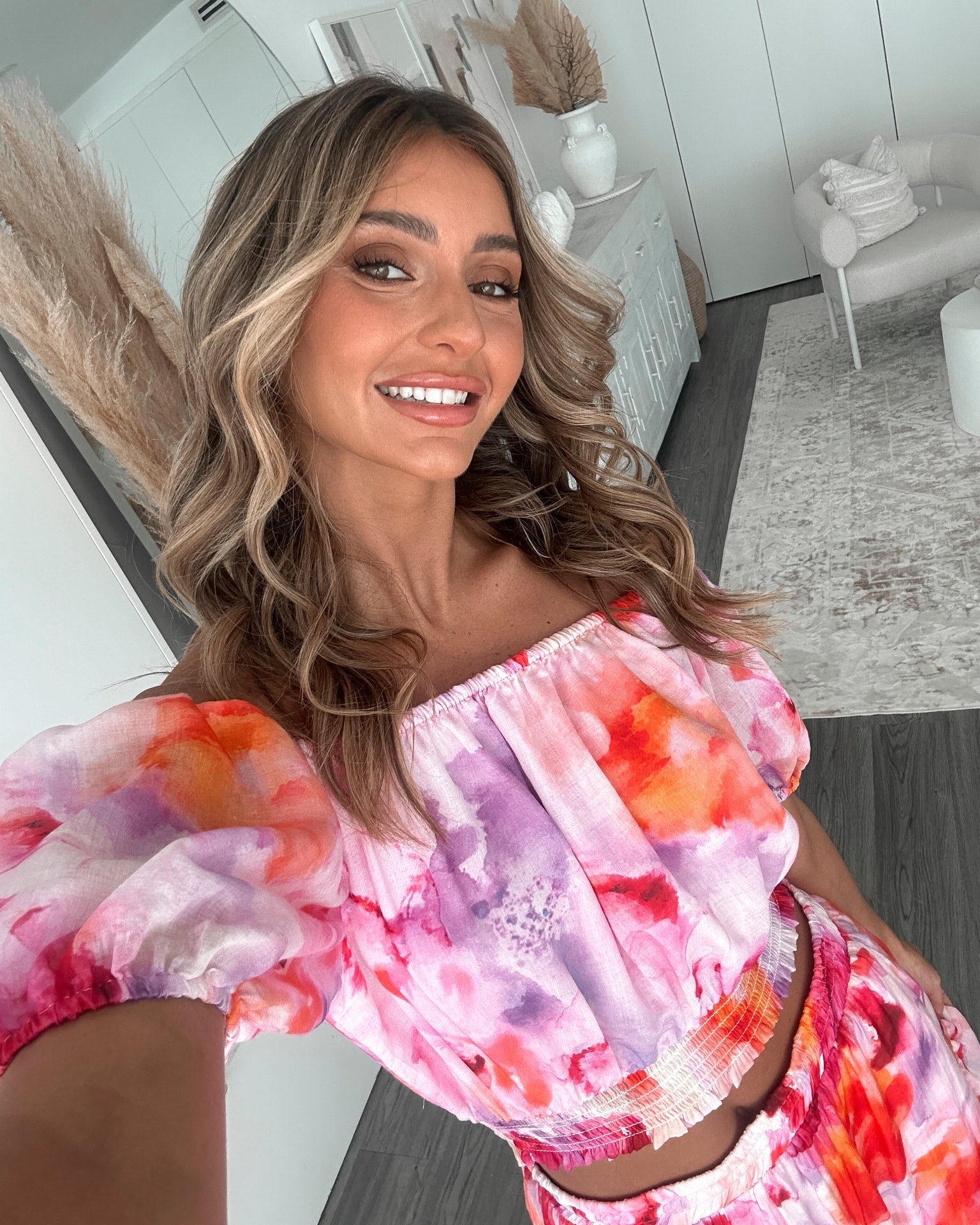 A woman stands indoors in a well-lit, modern room with sheer white curtains and minimalist white cabinetry. She is wearing a colorful off-the-shoulder two-piece outfit consisting of a ruffled crop top and a flowing maxi skirt with a vibrant watercolor floral print in pink, purple, orange, and red hues. Her wavy hair is styled loosely, and she stands gracefully with her hands gently clasped in front, exuding a fresh, summery elegance.