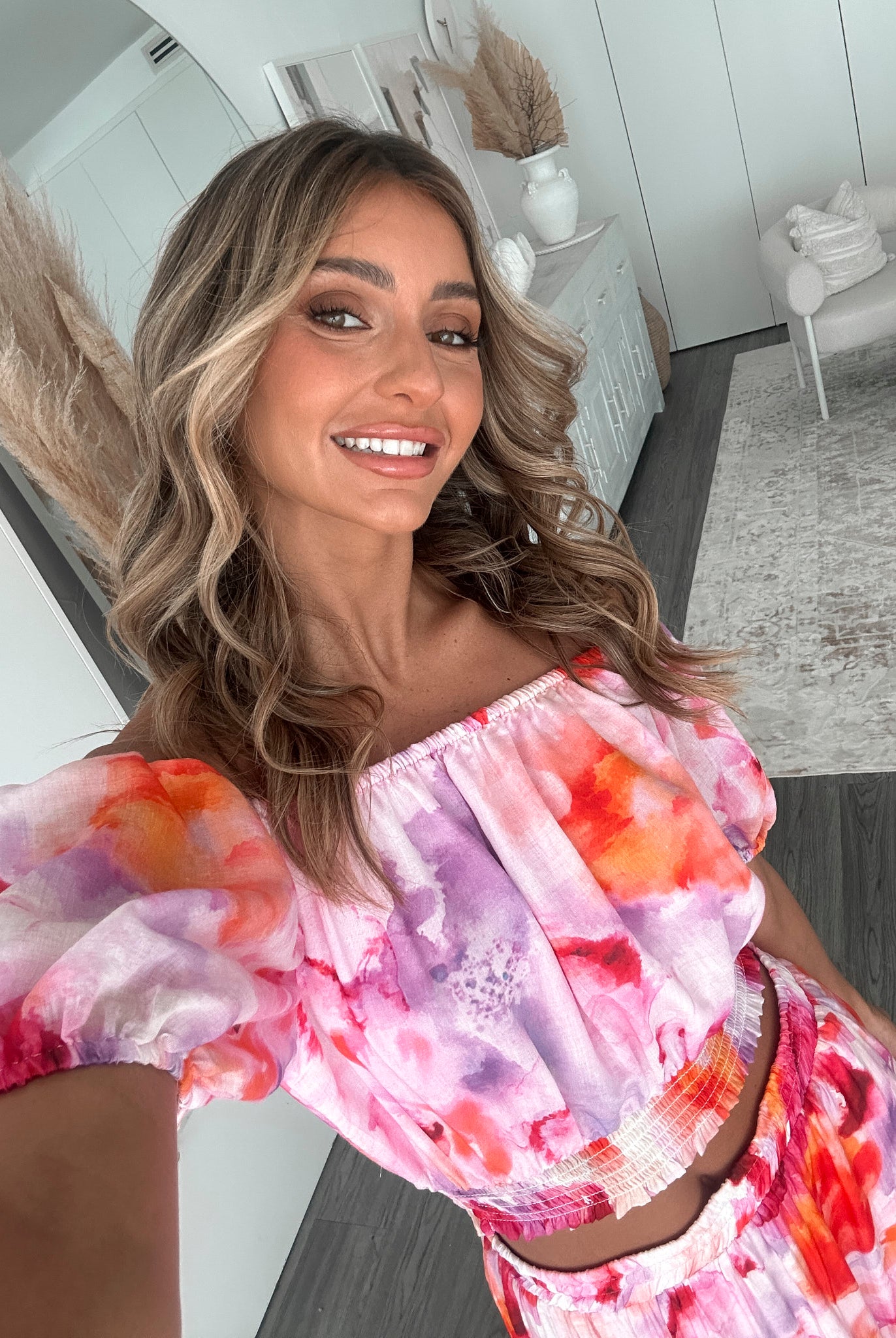 A woman stands indoors in a well-lit, modern room with sheer white curtains and minimalist white cabinetry. She is wearing a colorful off-the-shoulder two-piece outfit consisting of a ruffled crop top and a flowing maxi skirt with a vibrant watercolor floral print in pink, purple, orange, and red hues. Her wavy hair is styled loosely, and she stands gracefully with her hands gently clasped in front, exuding a fresh, summery elegance.