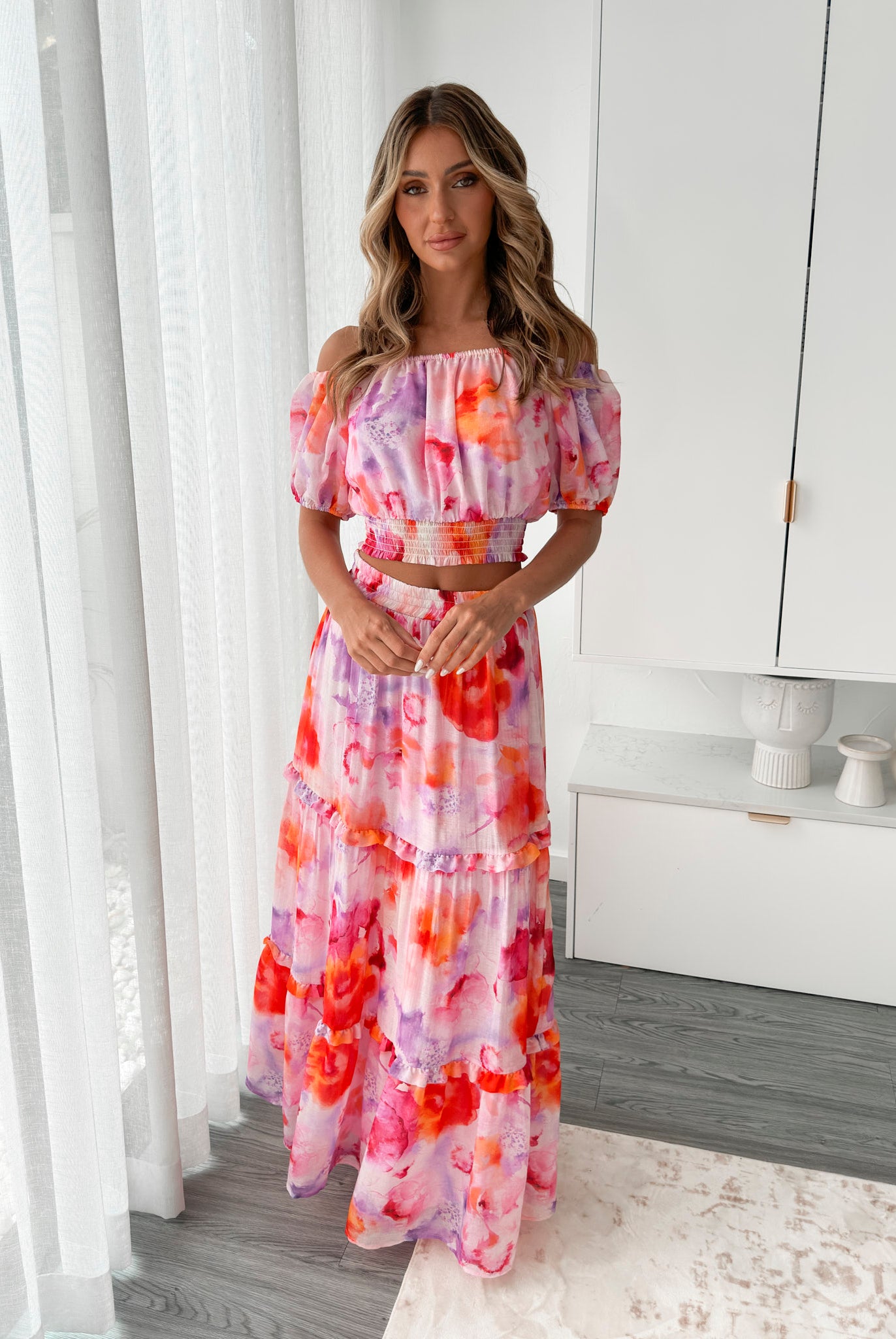 A woman stands indoors in a well-lit, modern room with sheer white curtains and minimalist white cabinetry. She is wearing a colorful off-the-shoulder two-piece outfit consisting of a ruffled crop top and a flowing maxi skirt with a vibrant watercolor floral print in pink, purple, orange, and red hues. Her wavy hair is styled loosely, and she stands gracefully with her hands gently clasped in front, exuding a fresh, summery elegance.