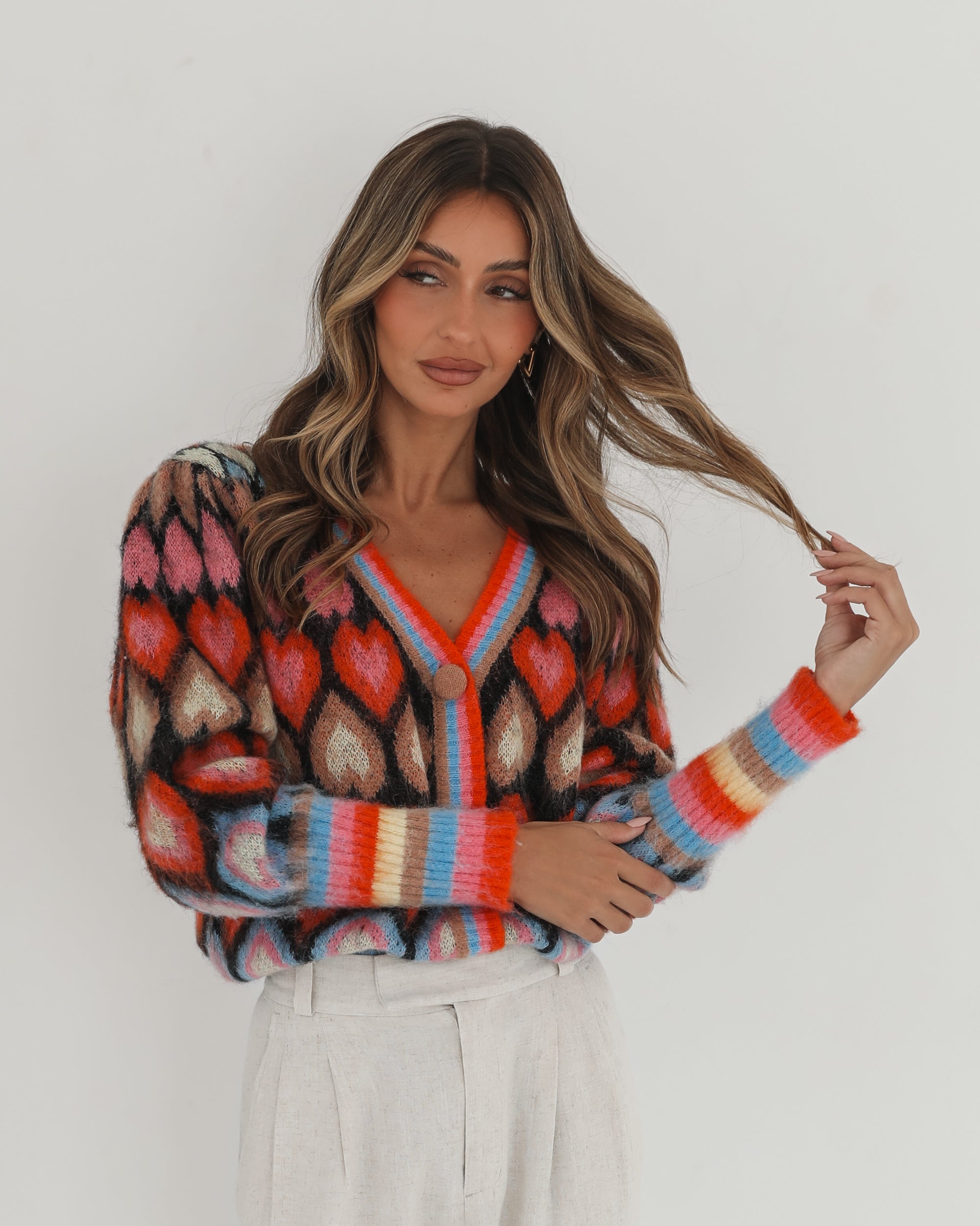 A woman poses against a plain white background wearing a colorful, patterned cardigan with heart shapes and bold stripes in red, pink, blue, and orange tones. She pairs it with high-waisted, light beige trousers and has long, wavy brown hair with blonde highlights. She is looking to the side and gently touching her hair with one hand.