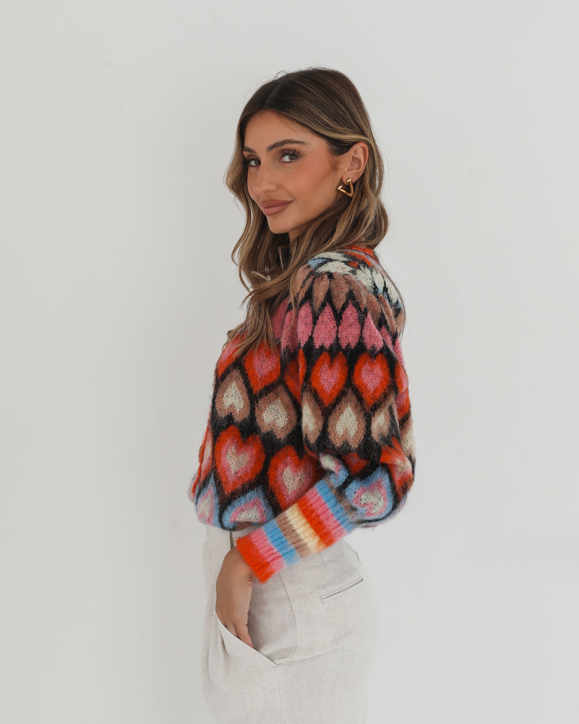 A woman poses against a plain white background wearing a colorful, patterned cardigan with heart shapes and bold stripes in red, pink, blue, and orange tones. She pairs it with high-waisted, light beige trousers and has long, wavy brown hair with blonde highlights. She is looking to the side and gently touching her hair with one hand.