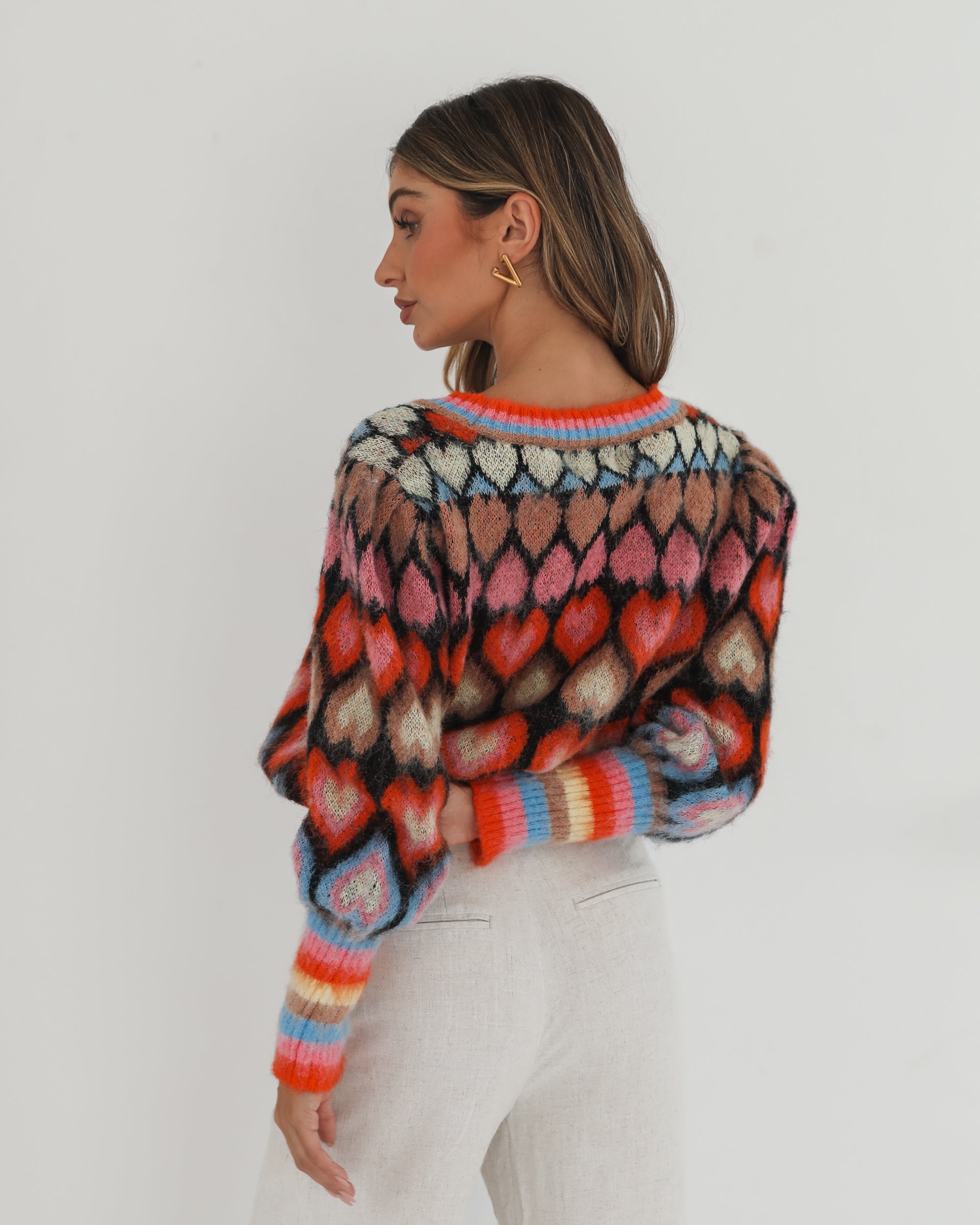 A woman poses against a plain white background wearing a colorful, patterned cardigan with heart shapes and bold stripes in red, pink, blue, and orange tones. She pairs it with high-waisted, light beige trousers and has long, wavy brown hair with blonde highlights. She is looking to the side and gently touching her hair with one hand.