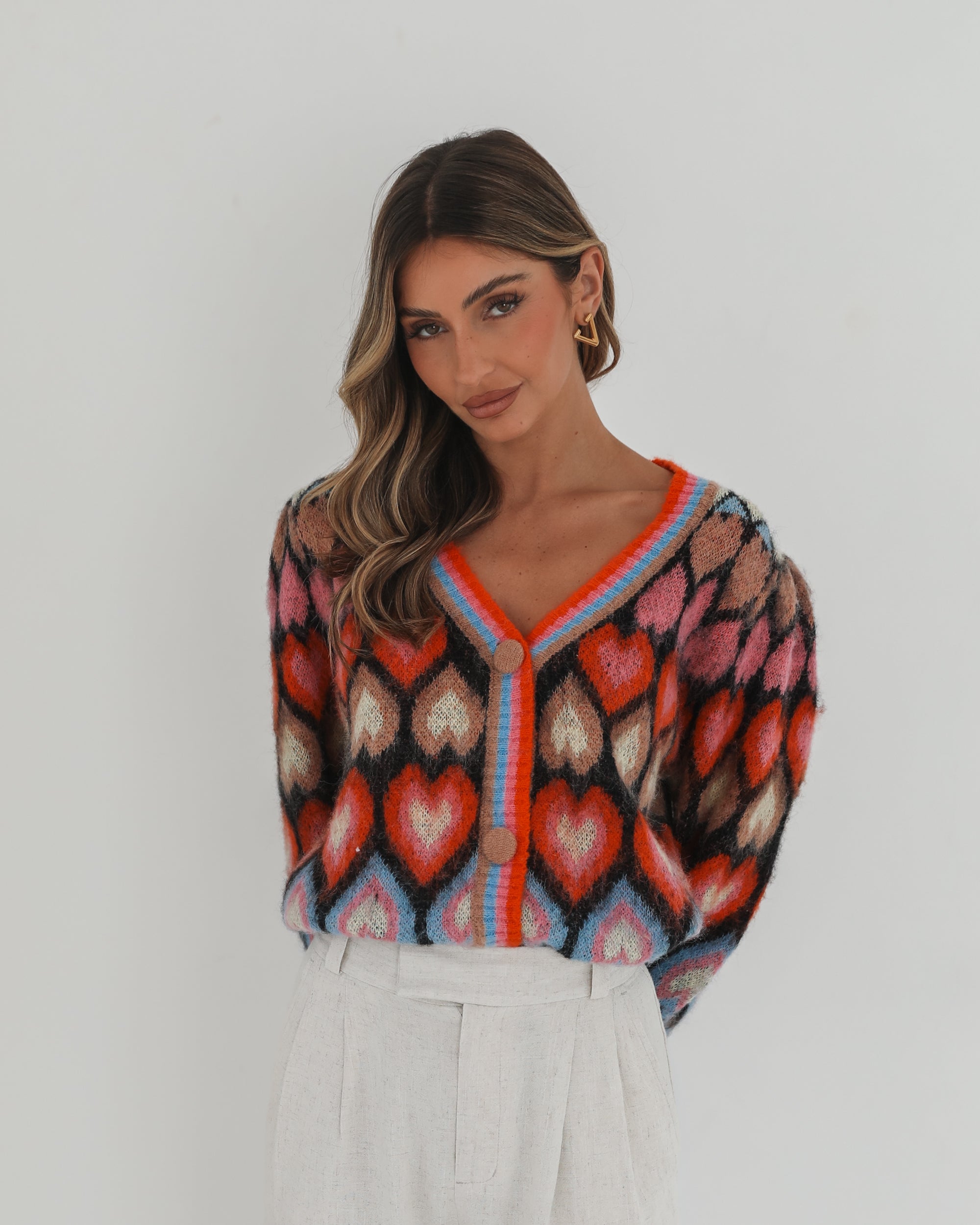 A woman poses against a plain white background wearing a colorful, patterned cardigan with heart shapes and bold stripes in red, pink, blue, and orange tones. She pairs it with high-waisted, light beige trousers and has long, wavy brown hair with blonde highlights. She is looking to the side and gently touching her hair with one hand.