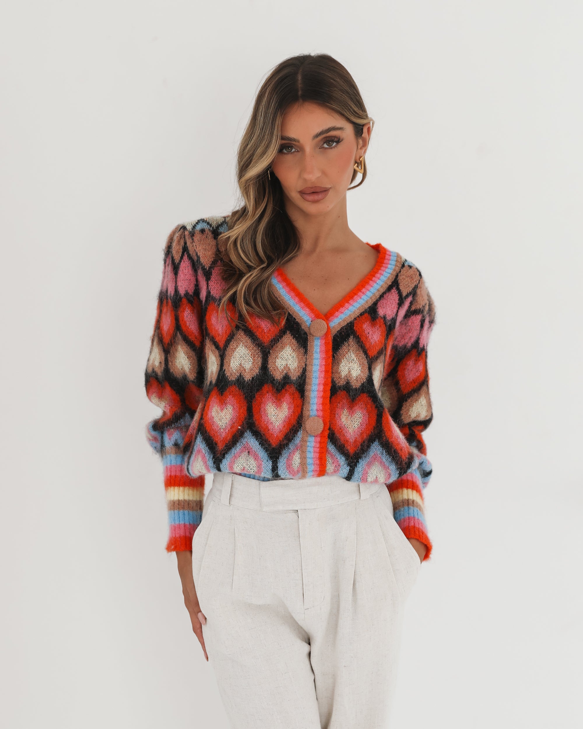 A woman poses against a plain white background wearing a colorful, patterned cardigan with heart shapes and bold stripes in red, pink, blue, and orange tones. She pairs it with high-waisted, light beige trousers and has long, wavy brown hair with blonde highlights. She is looking to the side and gently touching her hair with one hand.