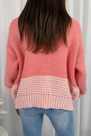 Person wearing a pink knitted sweater and jeans against a white curtain background
