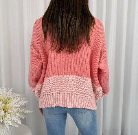 Person wearing a pink knitted sweater and jeans against a white curtain background