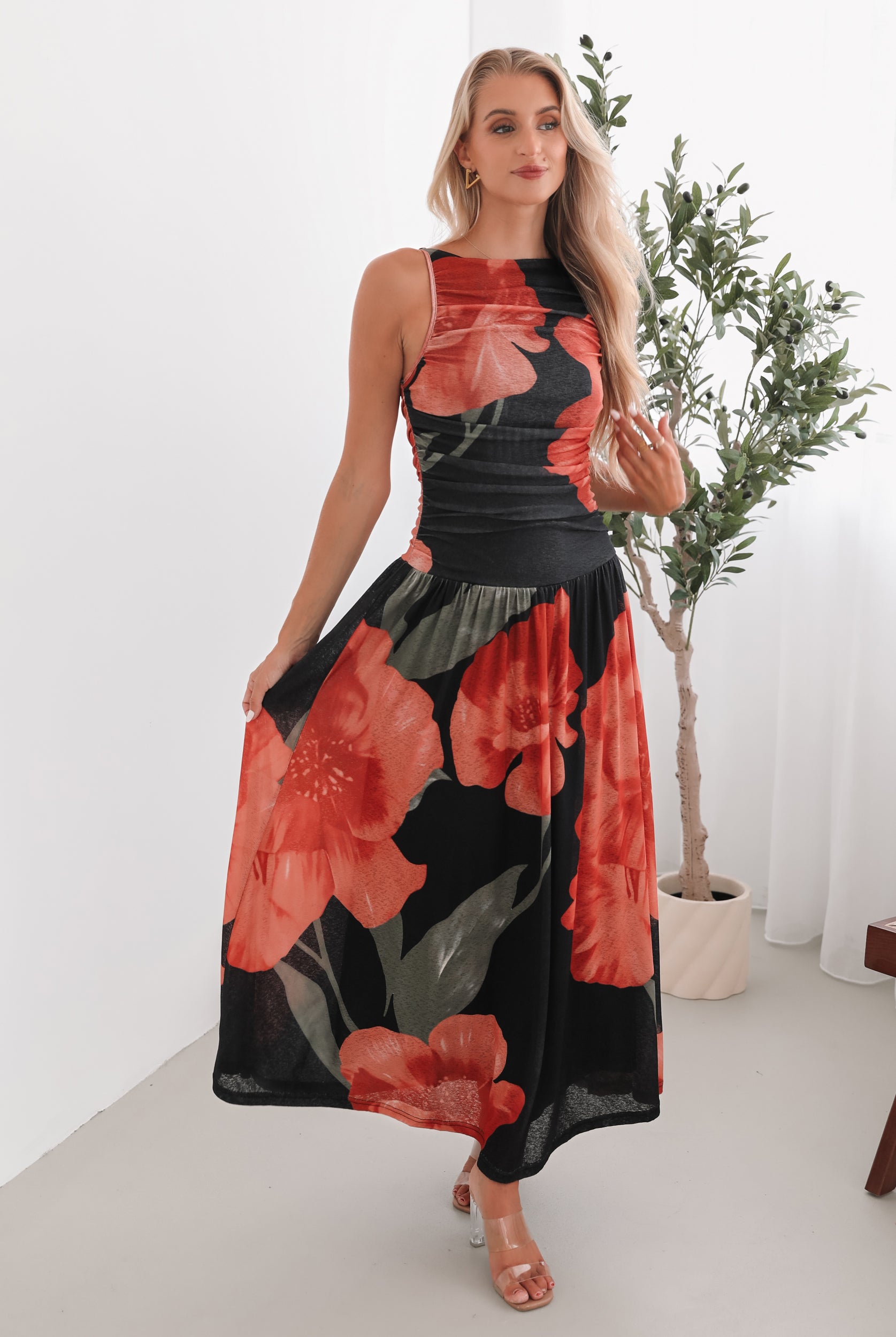A woman stands indoors near a potted plant, wearing a sleeveless black maxi dress with a bold red floral print. Her long blonde hair is styled in loose waves, and she accessorizes with gold hoop earrings and clear-heeled sandals. She smiles softly while posing with her hands gently holding the sides of her dress.
