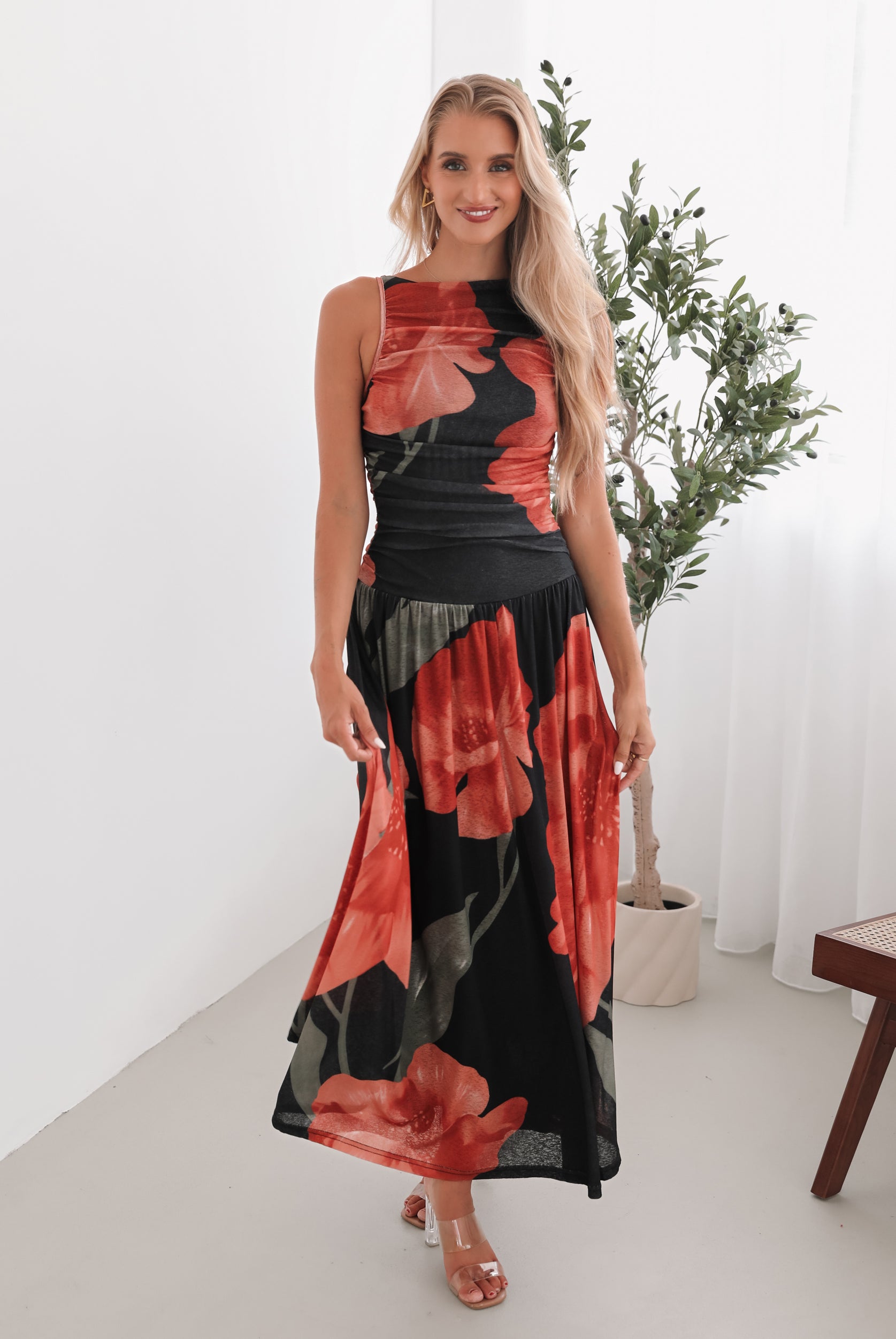 A woman stands indoors near a potted plant, wearing a sleeveless black maxi dress with a bold red floral print. Her long blonde hair is styled in loose waves, and she accessorizes with gold hoop earrings and clear-heeled sandals. She smiles softly while posing with her hands gently holding the sides of her dress.
