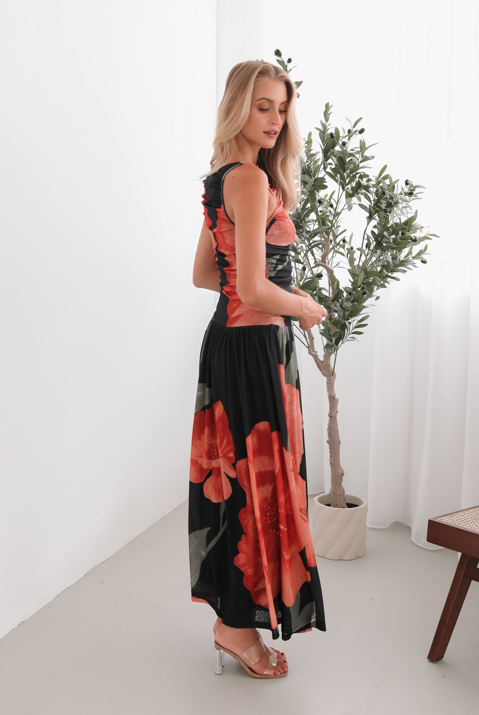 A woman stands indoors near a potted plant, wearing a sleeveless black maxi dress with a bold red floral print. Her long blonde hair is styled in loose waves, and she accessorizes with gold hoop earrings and clear-heeled sandals. She smiles softly while posing with her hands gently holding the sides of her dress.