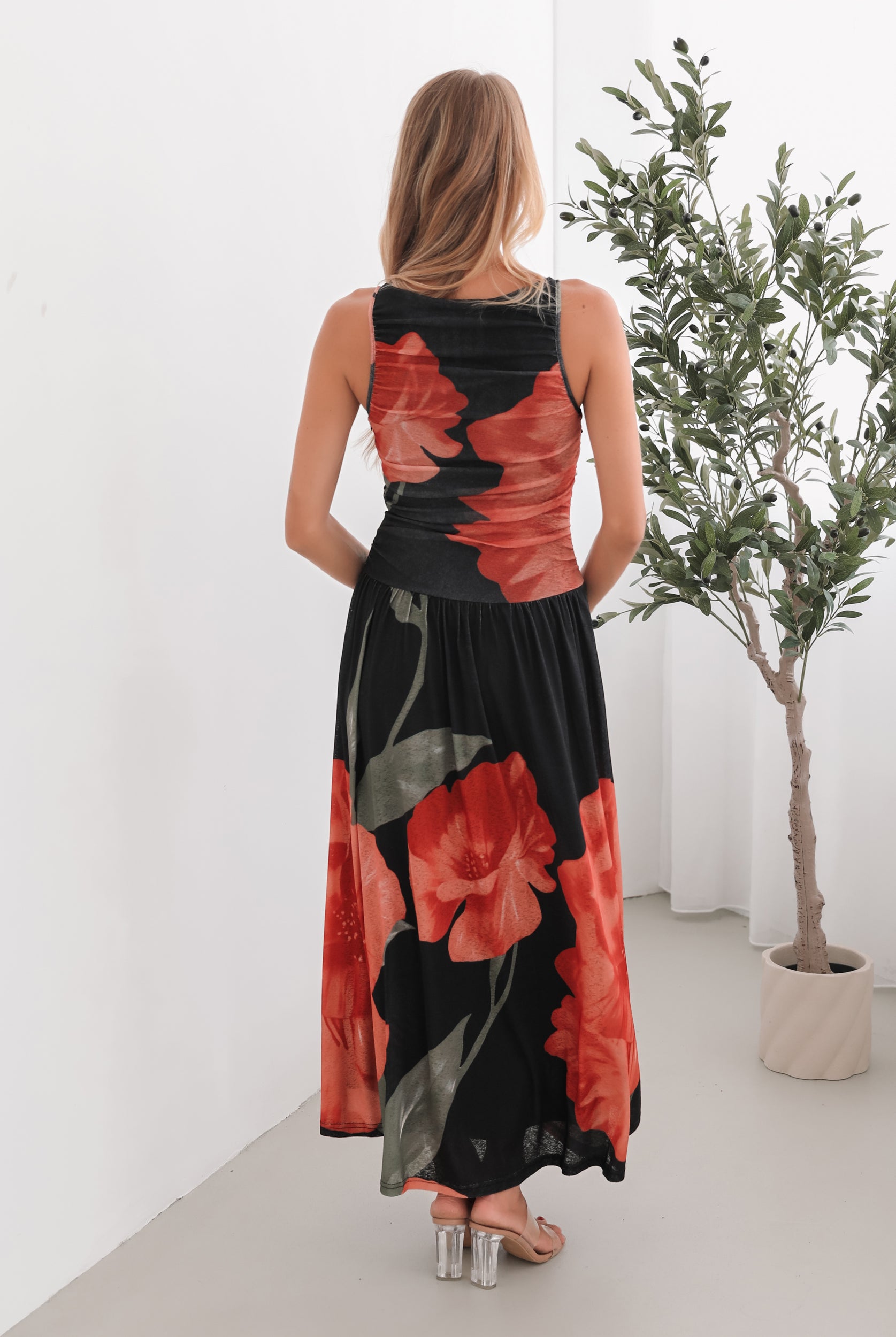 A woman stands indoors near a potted plant, wearing a sleeveless black maxi dress with a bold red floral print. Her long blonde hair is styled in loose waves, and she accessorizes with gold hoop earrings and clear-heeled sandals. She smiles softly while posing with her hands gently holding the sides of her dress.