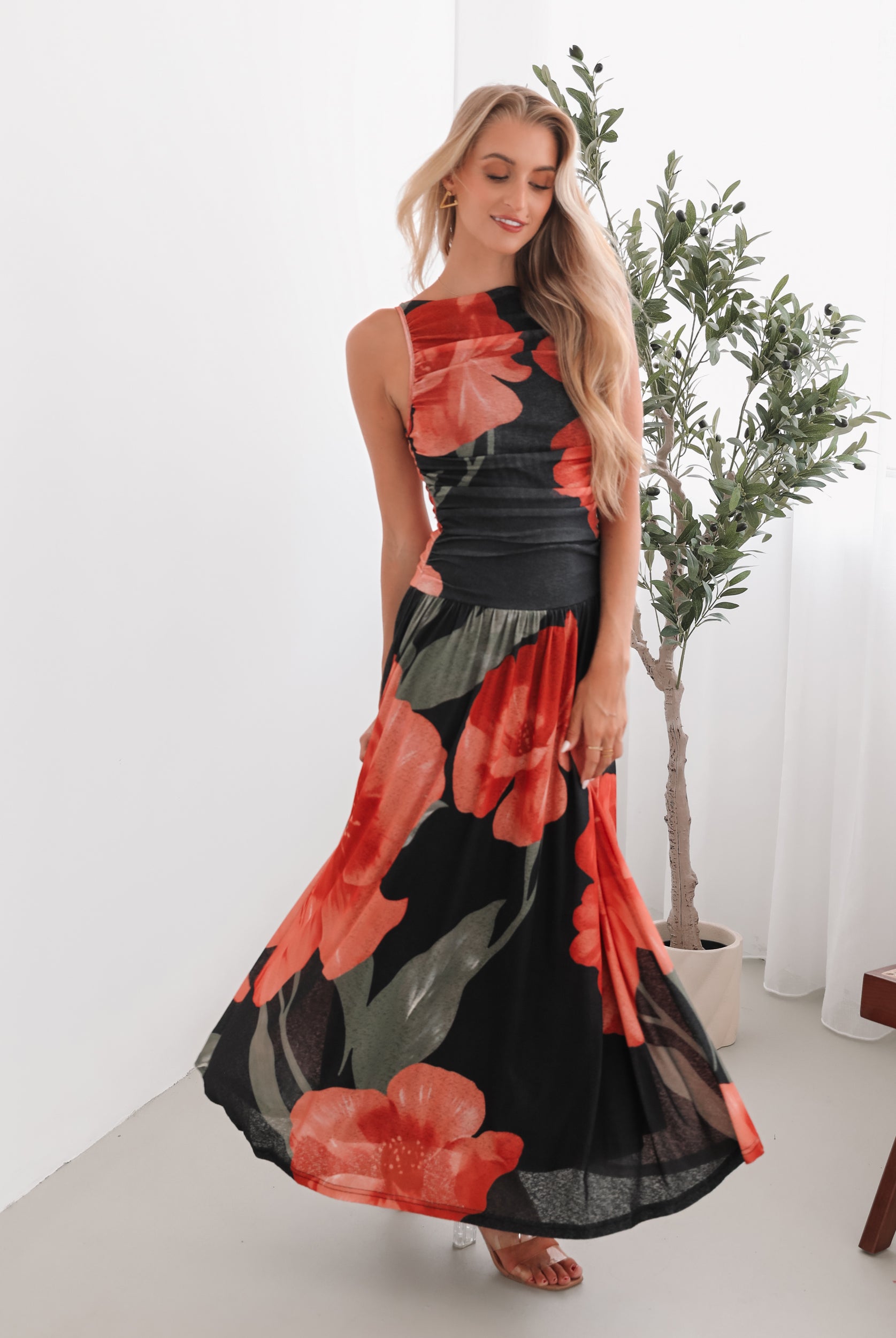 A woman stands indoors near a potted plant, wearing a sleeveless black maxi dress with a bold red floral print. Her long blonde hair is styled in loose waves, and she accessorizes with gold hoop earrings and clear-heeled sandals. She smiles softly while posing with her hands gently holding the sides of her dress.