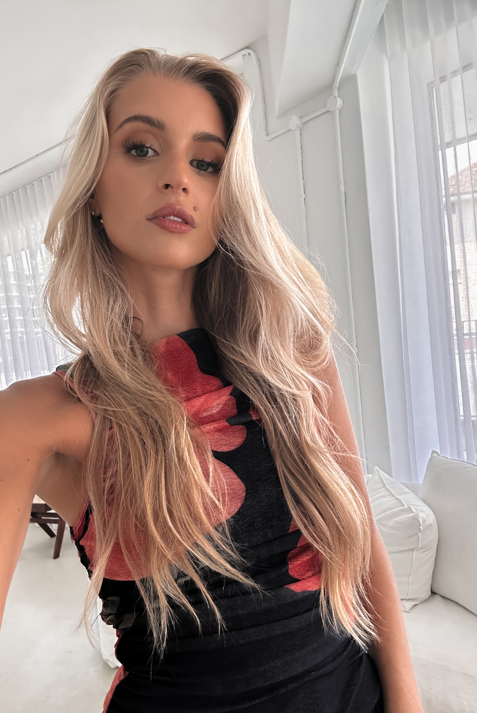 A woman stands indoors near a potted plant, wearing a sleeveless black maxi dress with a bold red floral print. Her long blonde hair is styled in loose waves, and she accessorizes with gold hoop earrings and clear-heeled sandals. She smiles softly while posing with her hands gently holding the sides of her dress.