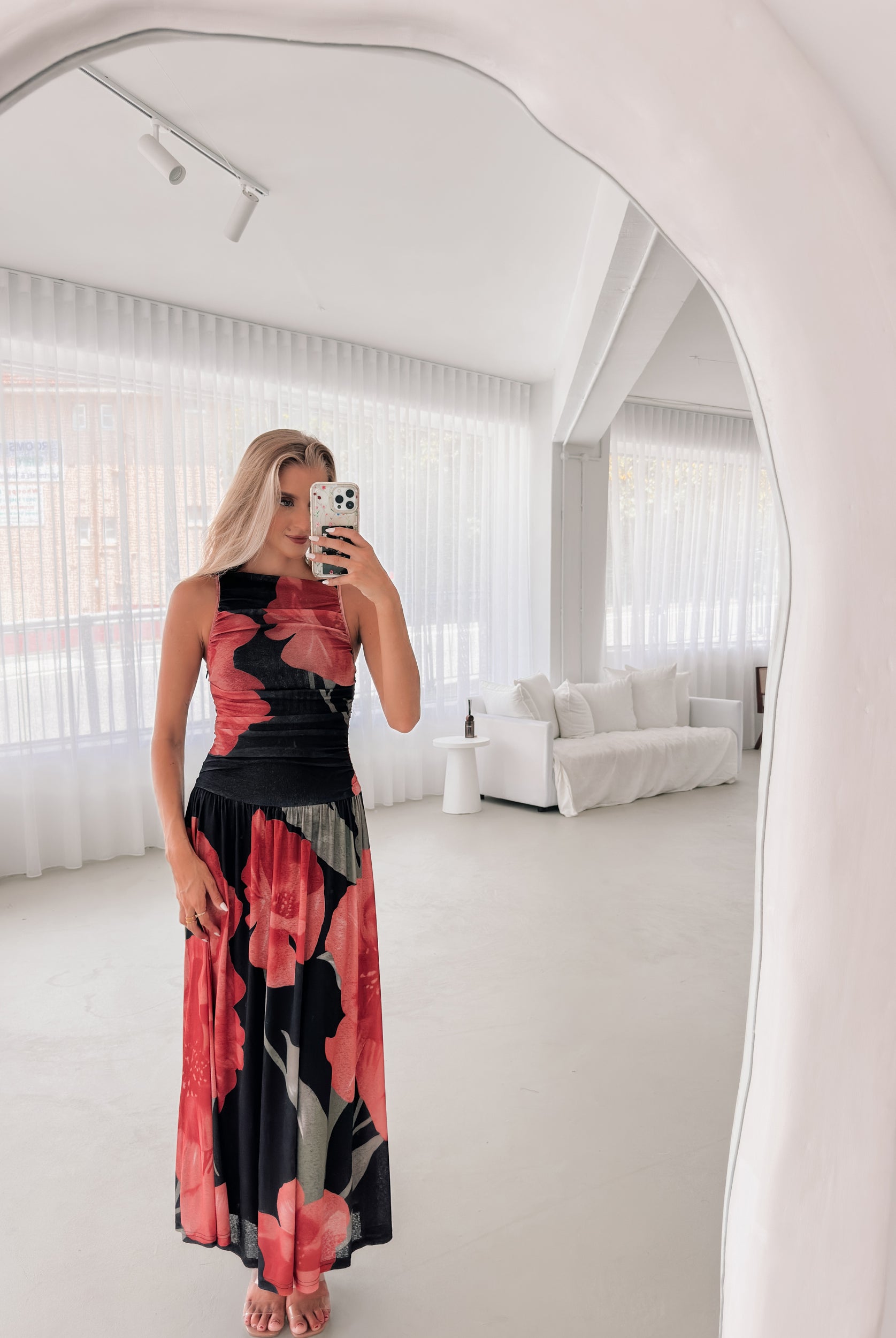 A woman stands indoors near a potted plant, wearing a sleeveless black maxi dress with a bold red floral print. Her long blonde hair is styled in loose waves, and she accessorizes with gold hoop earrings and clear-heeled sandals. She smiles softly while posing with her hands gently holding the sides of her dress.