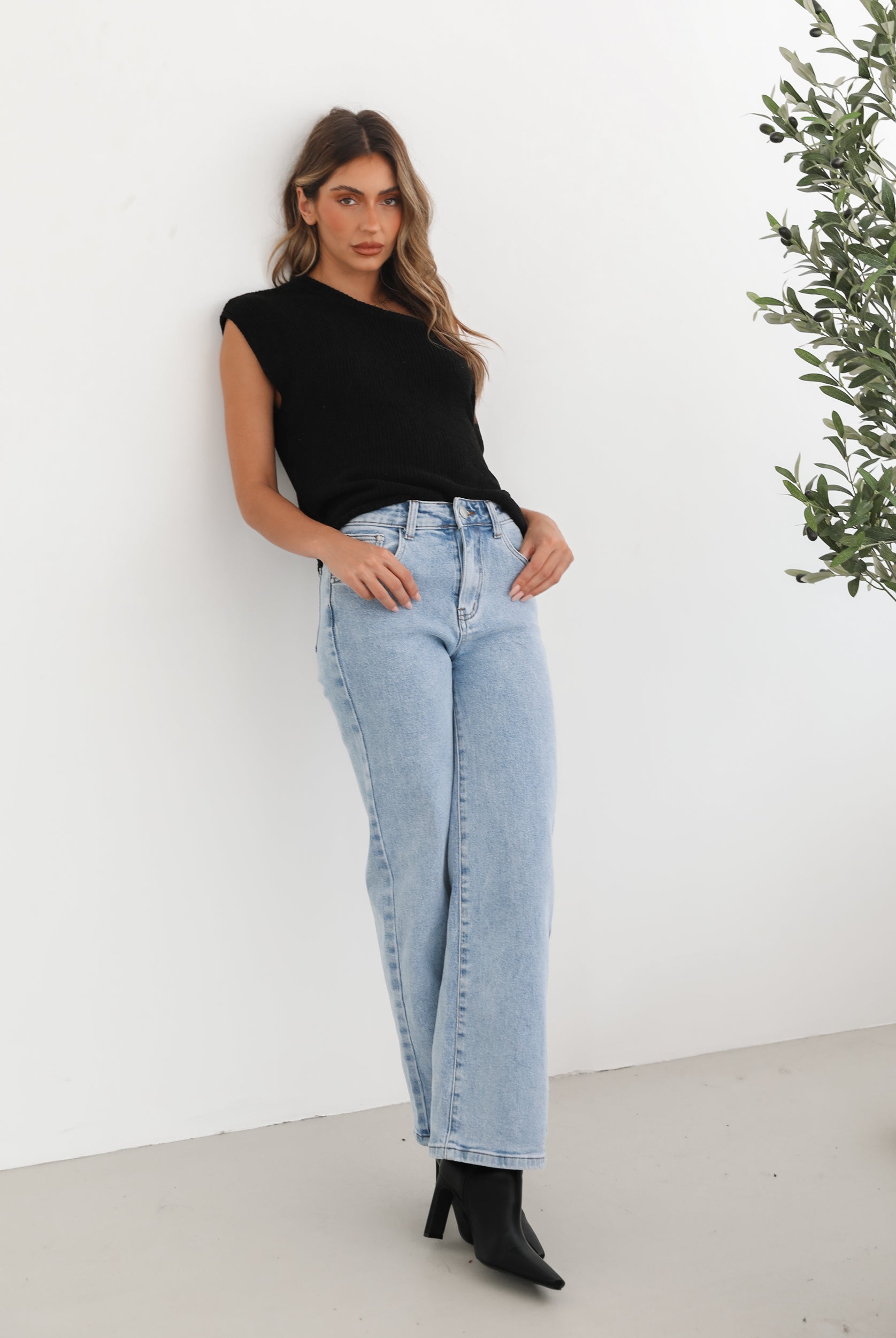 Woman wearing light-wash flared jeans and black heeled ankle boots, paired with a sleeveless black top, standing indoors near a potted plant and a wooden chair.