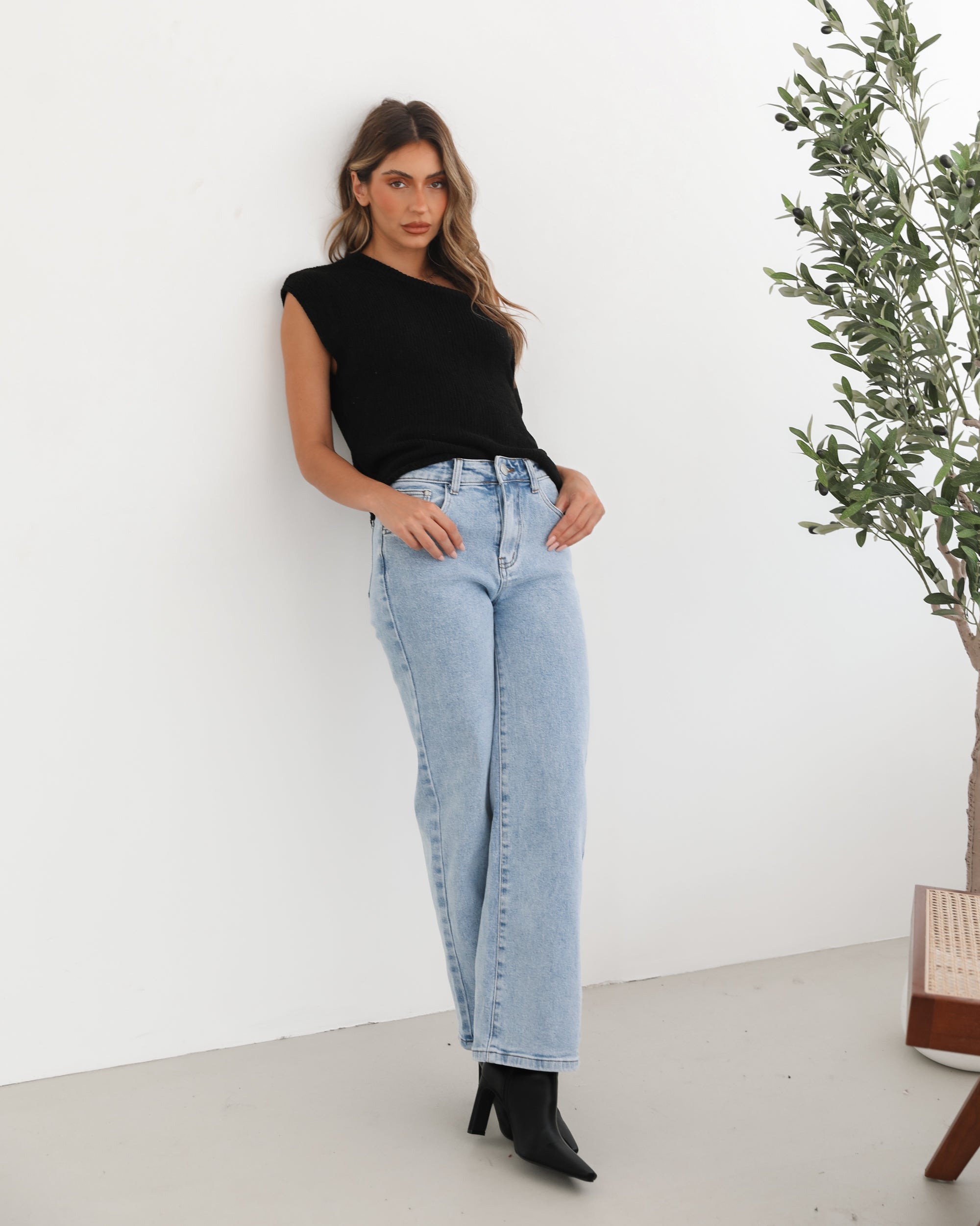 Woman wearing light-wash flared jeans and black heeled ankle boots, paired with a sleeveless black top, standing indoors near a potted plant and a wooden chair.