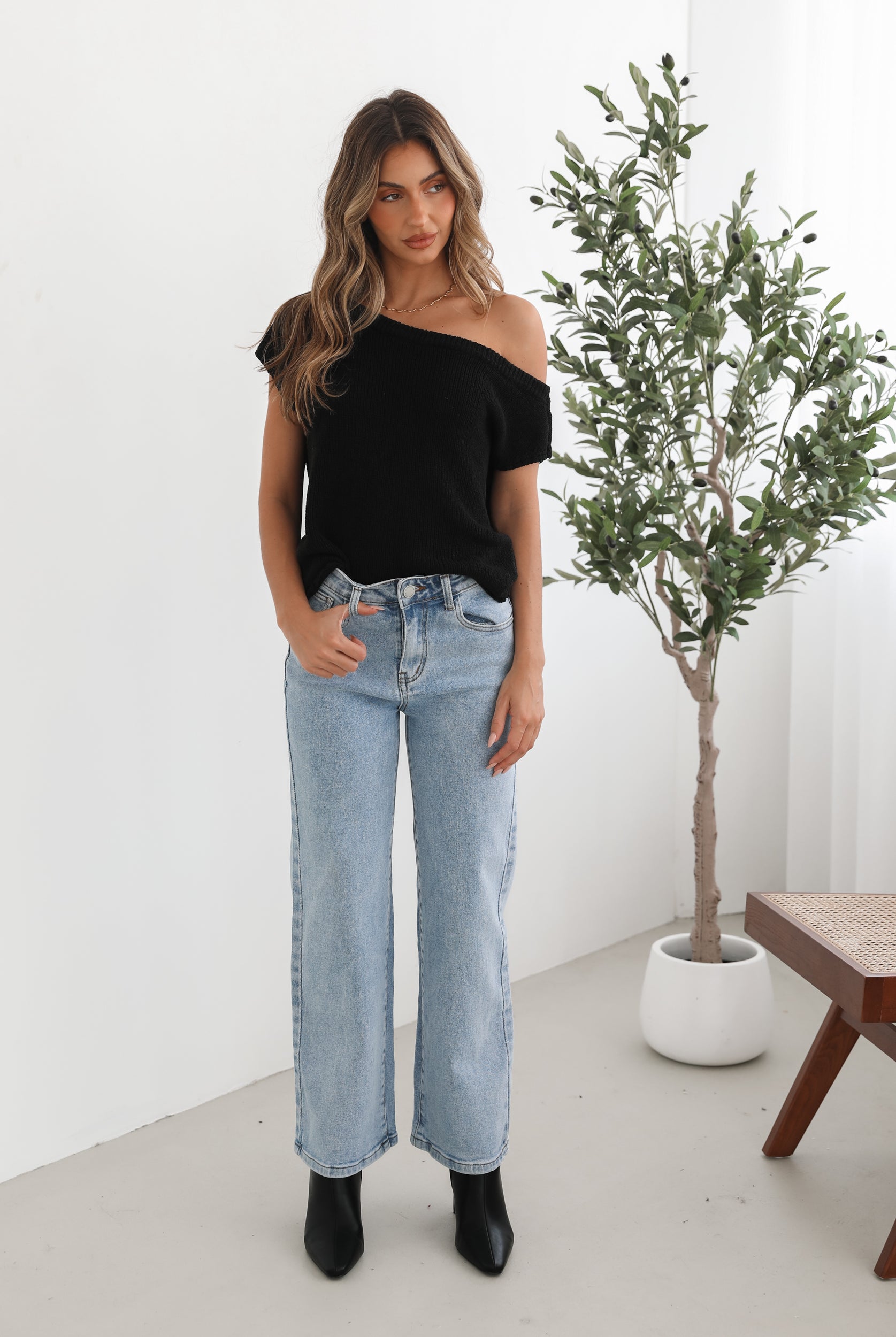 Woman wearing light-wash flared jeans and black heeled ankle boots, paired with a sleeveless black top, standing indoors near a potted plant and a wooden chair.