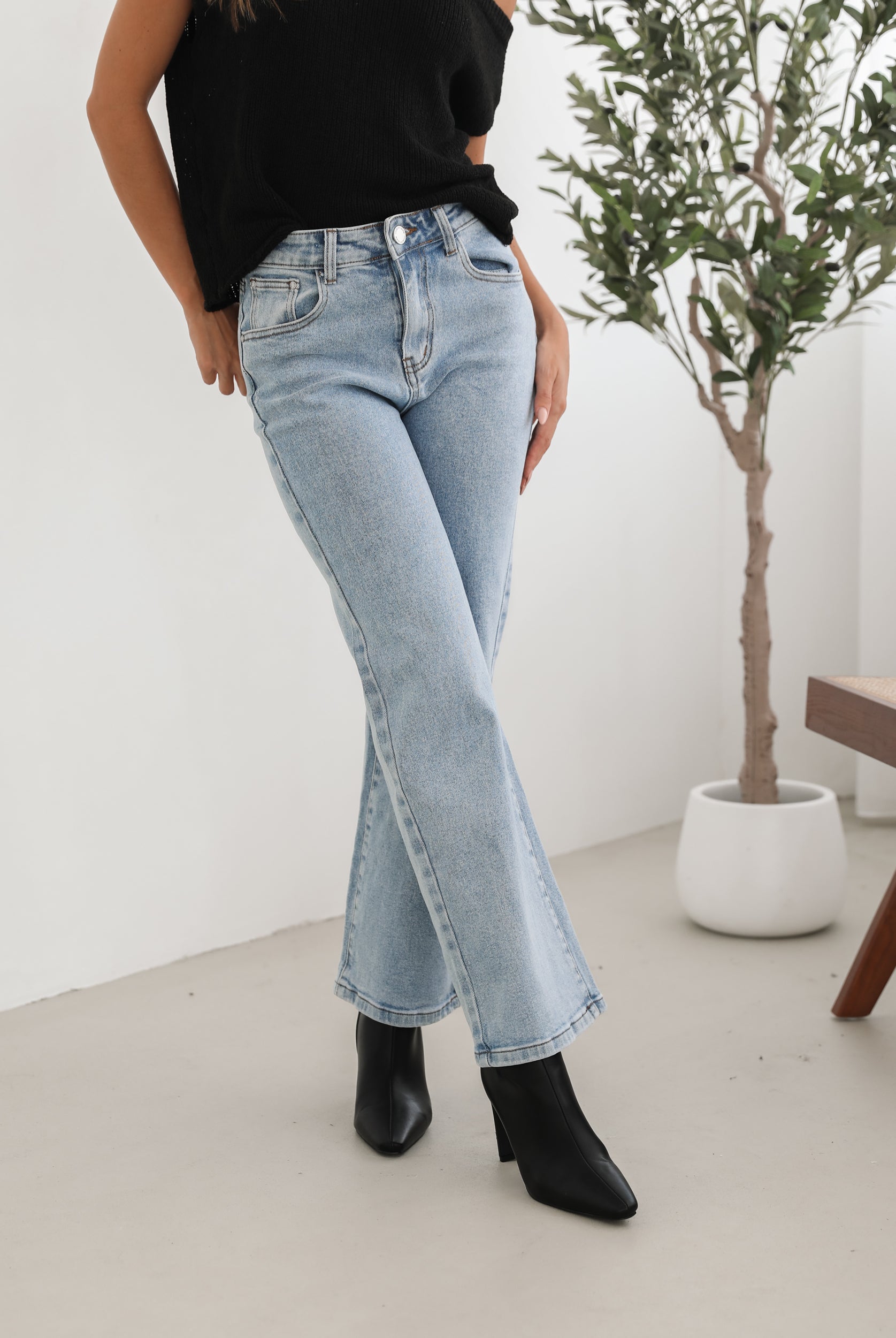 Woman wearing light-wash flared jeans and black heeled ankle boots, paired with a sleeveless black top, standing indoors near a potted plant and a wooden chair.