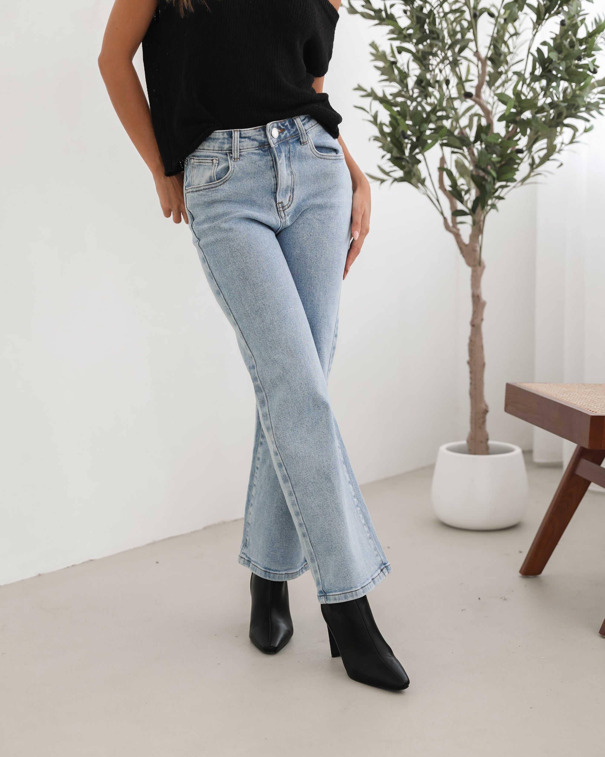 Woman wearing light-wash flared jeans and black heeled ankle boots, paired with a sleeveless black top, standing indoors near a potted plant and a wooden chair.