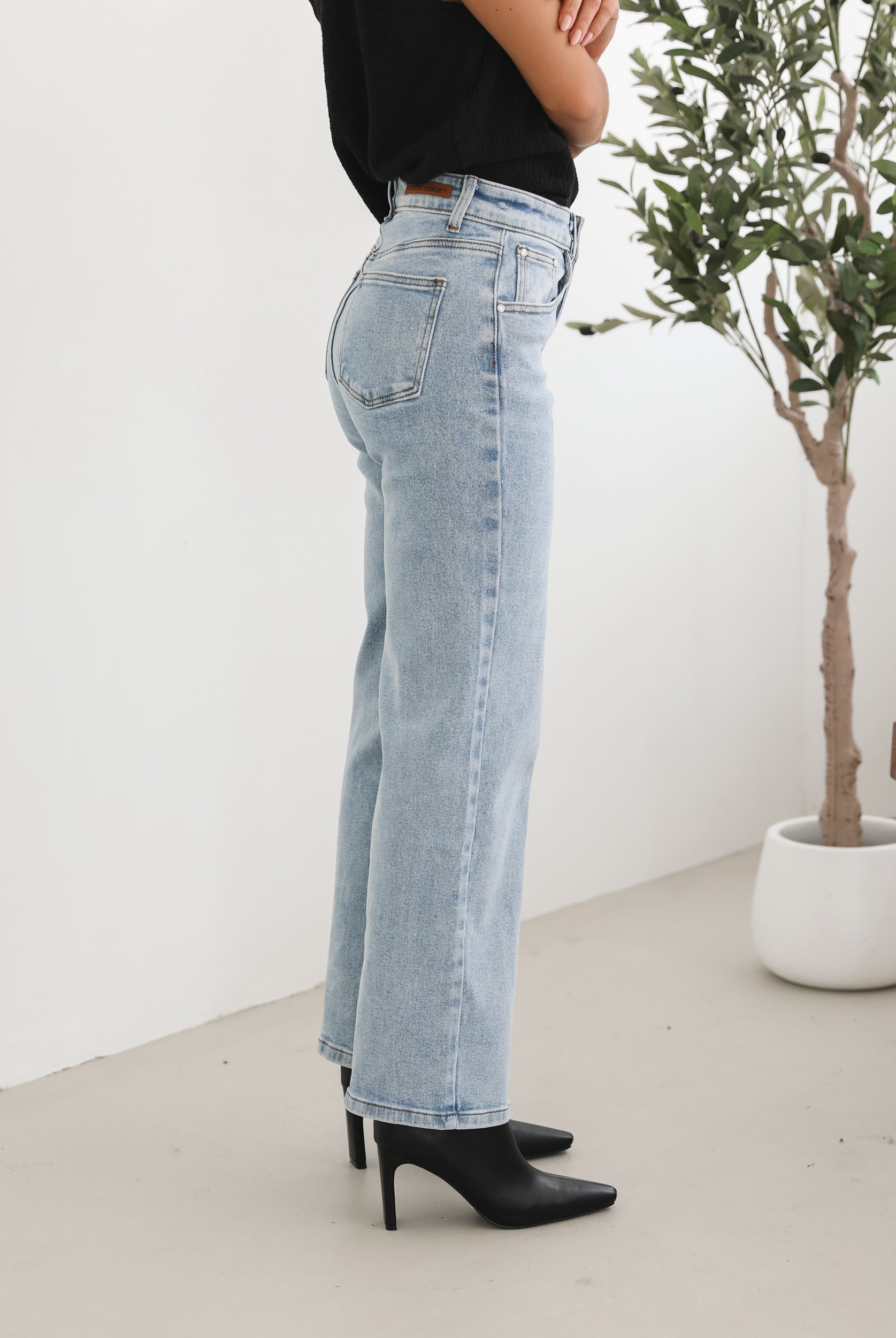 Woman wearing light-wash flared jeans and black heeled ankle boots, paired with a sleeveless black top, standing indoors near a potted plant and a wooden chair.