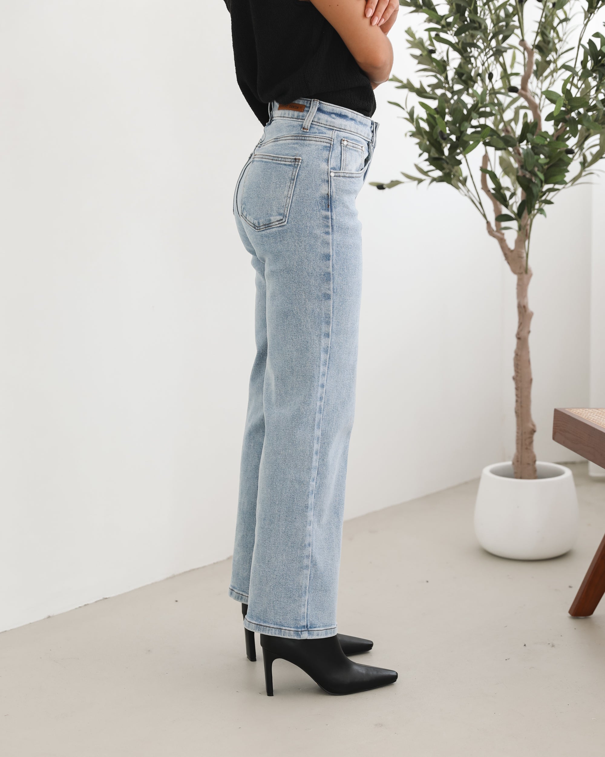 Woman wearing light-wash flared jeans and black heeled ankle boots, paired with a sleeveless black top, standing indoors near a potted plant and a wooden chair.