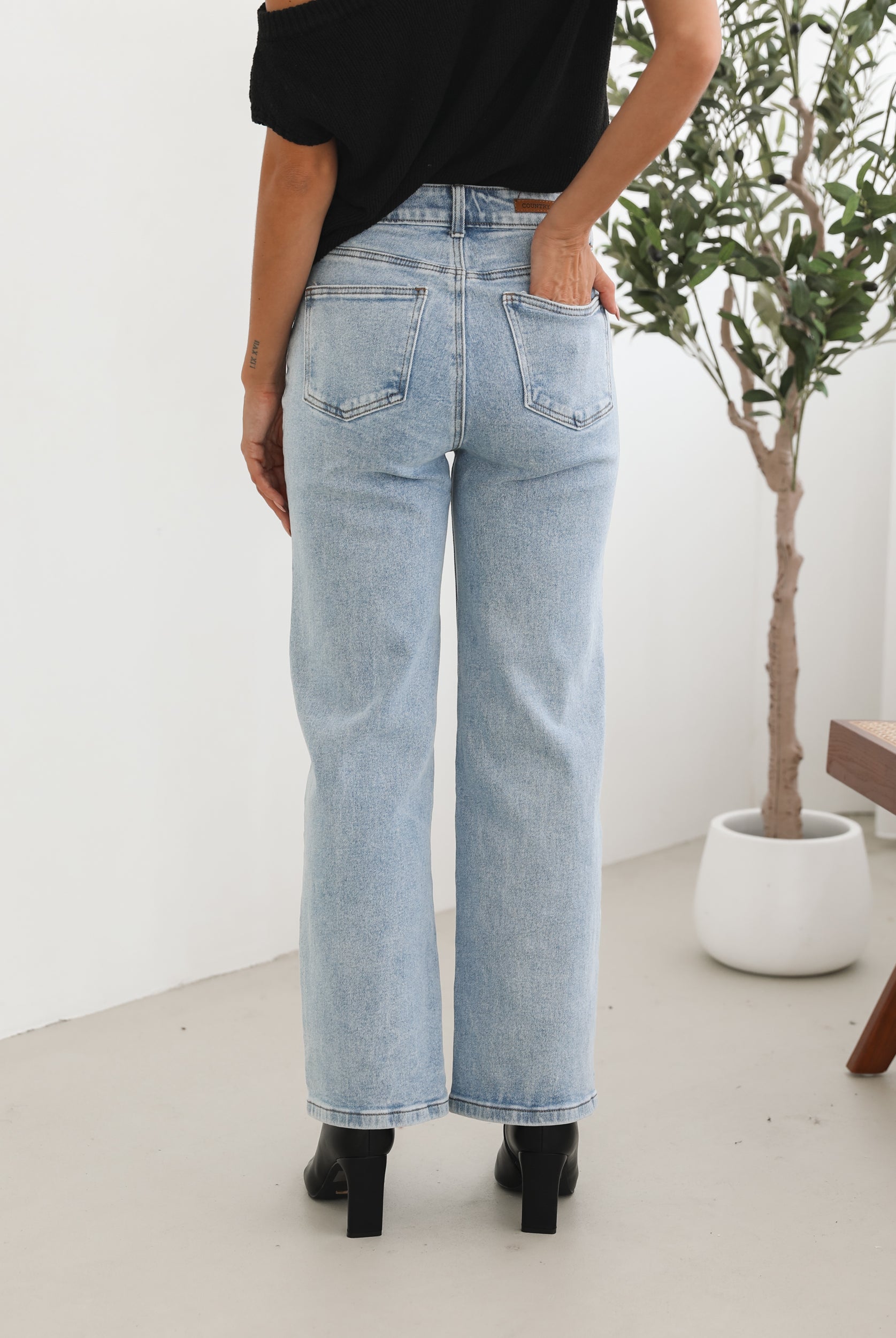 Woman wearing light-wash flared jeans and black heeled ankle boots, paired with a sleeveless black top, standing indoors near a potted plant and a wooden chair.
