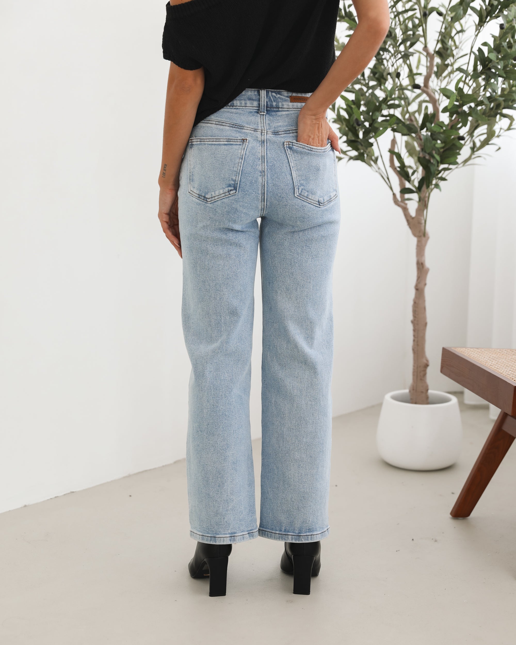 Woman wearing light-wash flared jeans and black heeled ankle boots, paired with a sleeveless black top, standing indoors near a potted plant and a wooden chair.
