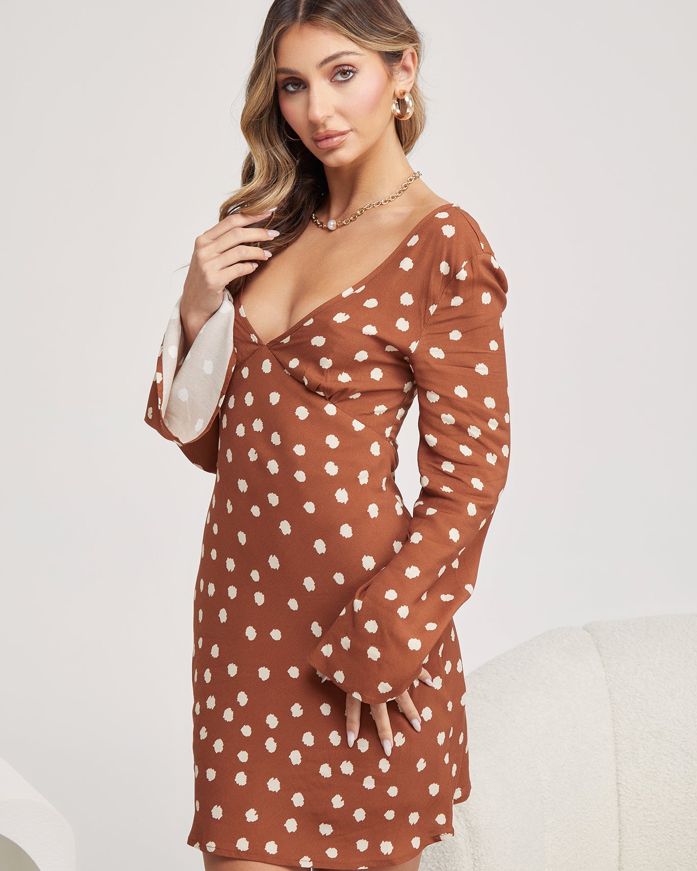 Woman wearing a brown long-sleeve mini dress with white polka dots and flared cuffs, paired with light beige ankle boots, standing in a modern, minimalist room with a white chair, a small shelf, and a textured rug.
