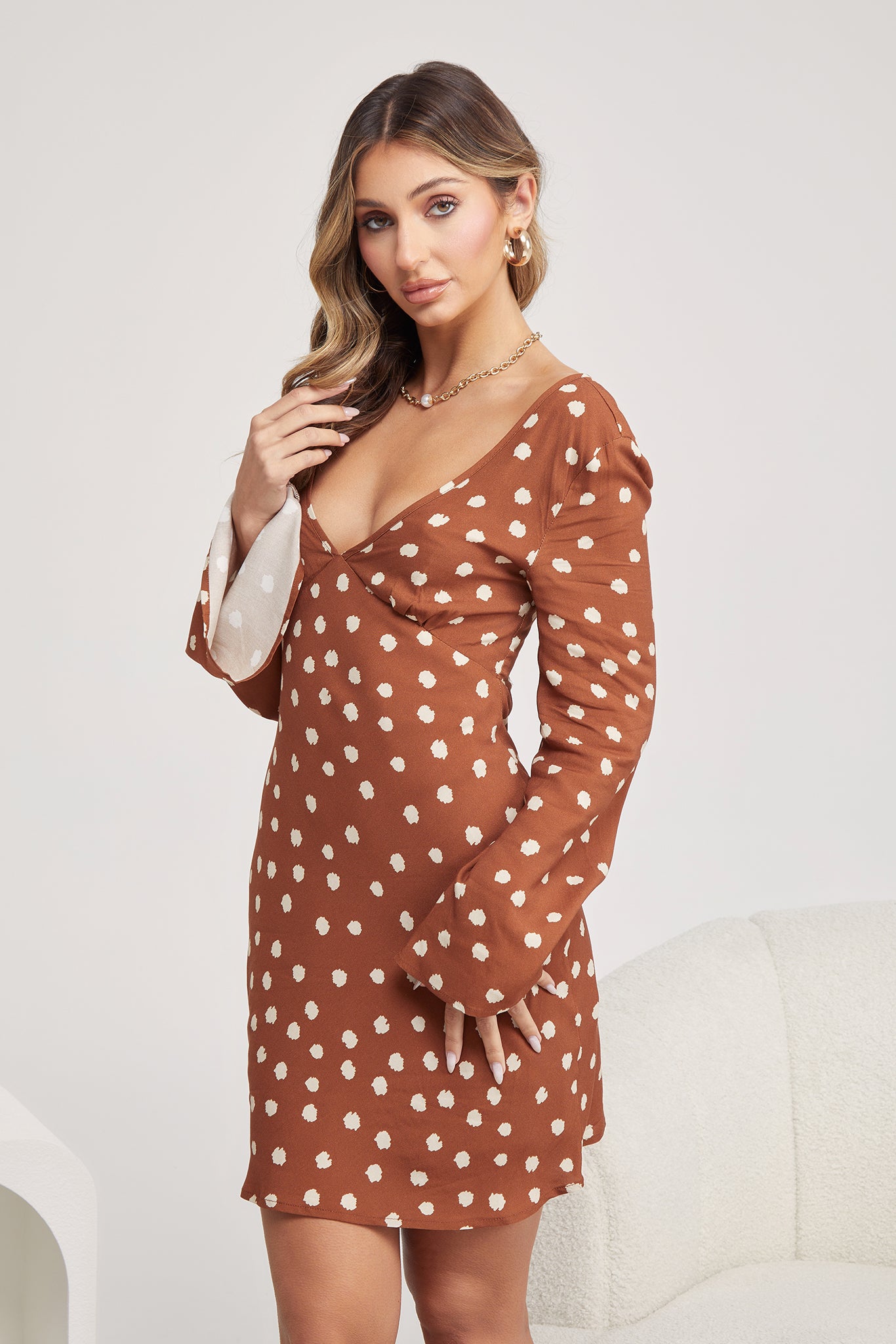 Woman wearing a brown long-sleeve mini dress with white polka dots and flared cuffs, paired with light beige ankle boots, standing in a modern, minimalist room with a white chair, a small shelf, and a textured rug.