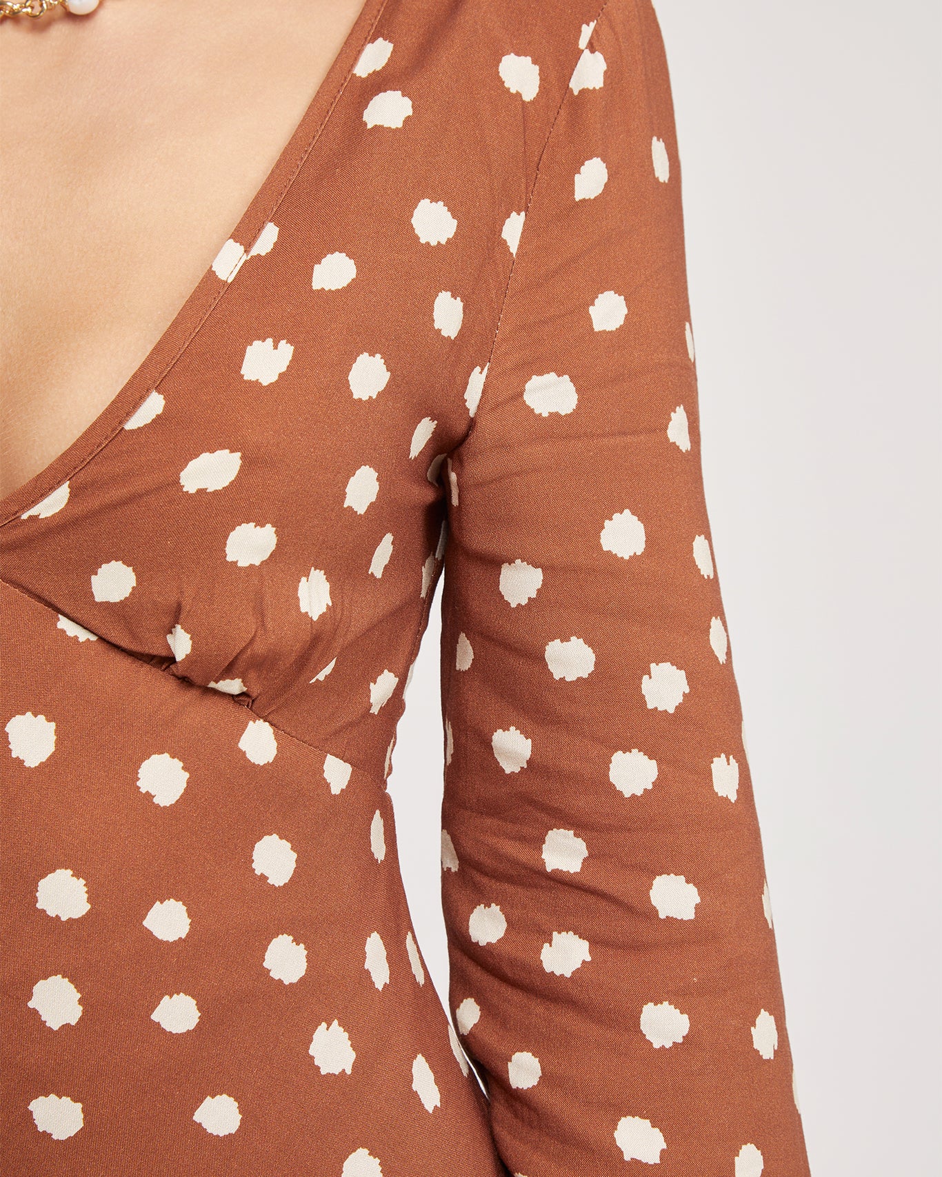 Woman wearing a brown long-sleeve mini dress with white polka dots and flared cuffs, paired with light beige ankle boots, standing in a modern, minimalist room with a white chair, a small shelf, and a textured rug.