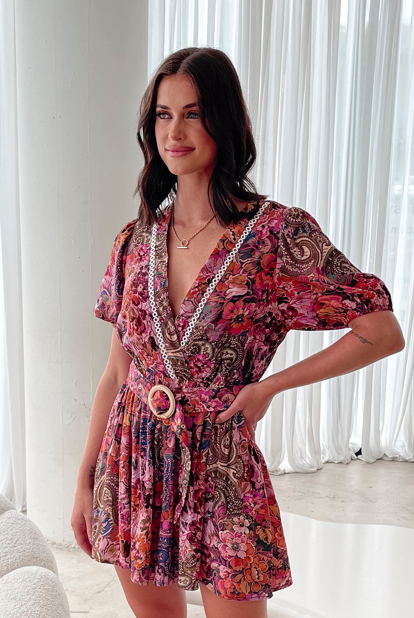 Woman wearing a short, colorful floral and paisley-patterned dress with a deep V-neckline trimmed in lace, puff sleeves, and a belted waist, standing indoors in front of sheer white curtains.