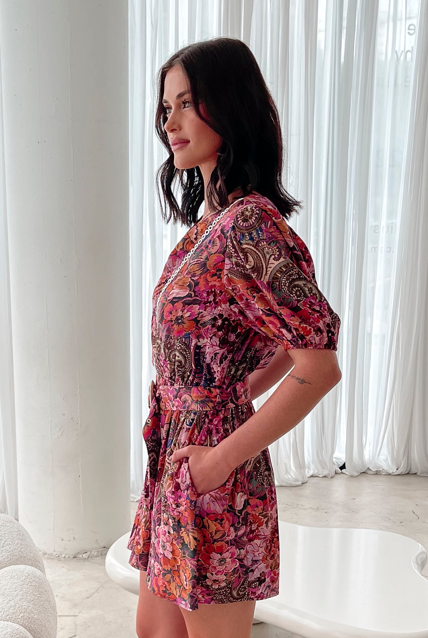 Woman wearing a short, colorful floral and paisley-patterned dress with a deep V-neckline trimmed in lace, puff sleeves, and a belted waist, standing indoors in front of sheer white curtains.