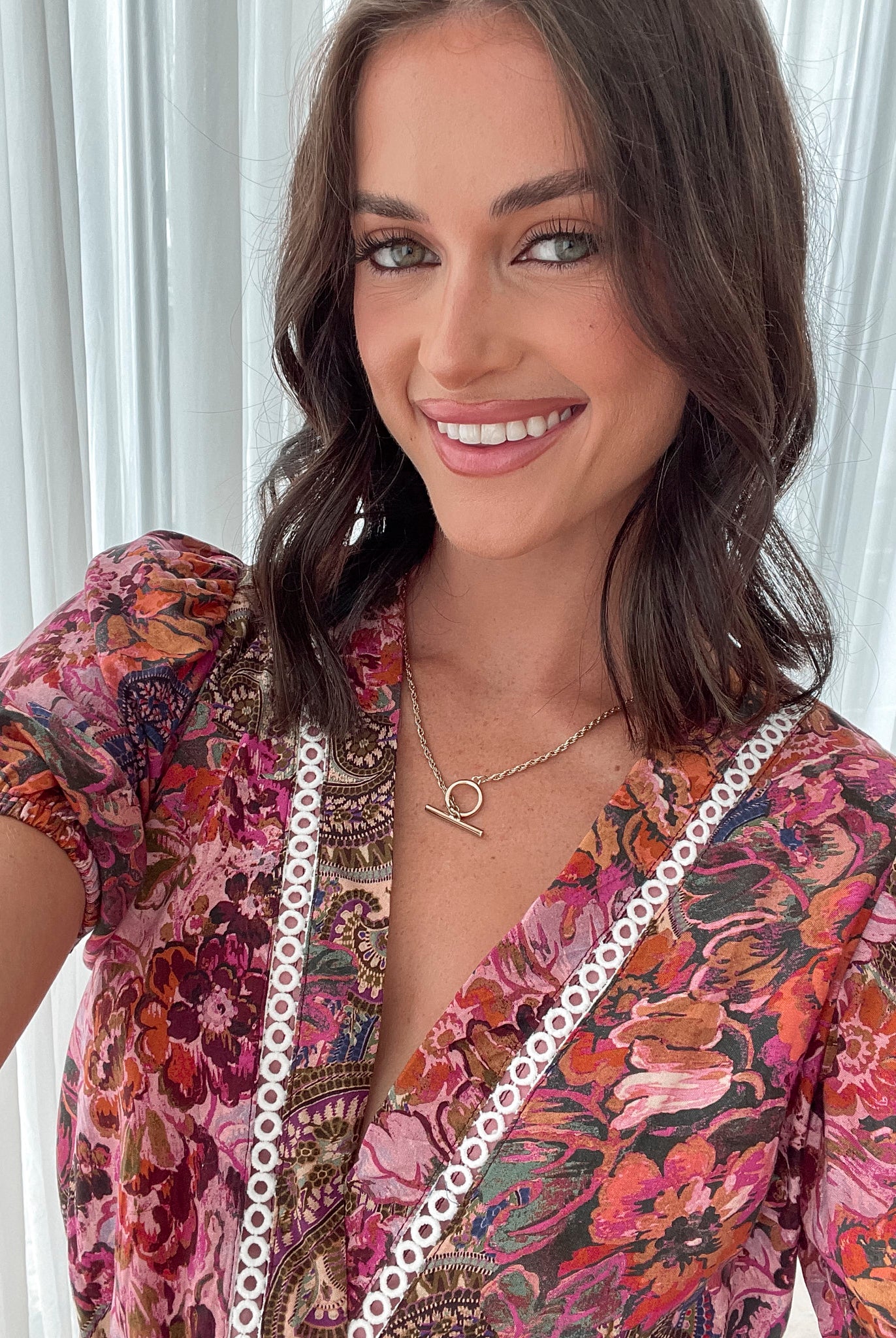 Woman wearing a short, colorful floral and paisley-patterned dress with a deep V-neckline trimmed in lace, puff sleeves, and a belted waist, standing indoors in front of sheer white curtains.