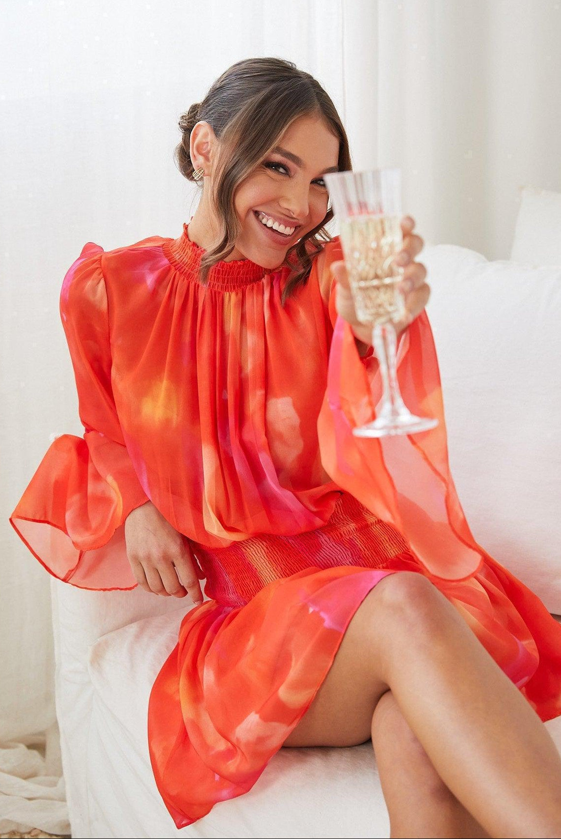 Woman sitting on white couch wearing the Daenerys Orange Print Mini Dress with high neckline and long trumpet sleeves