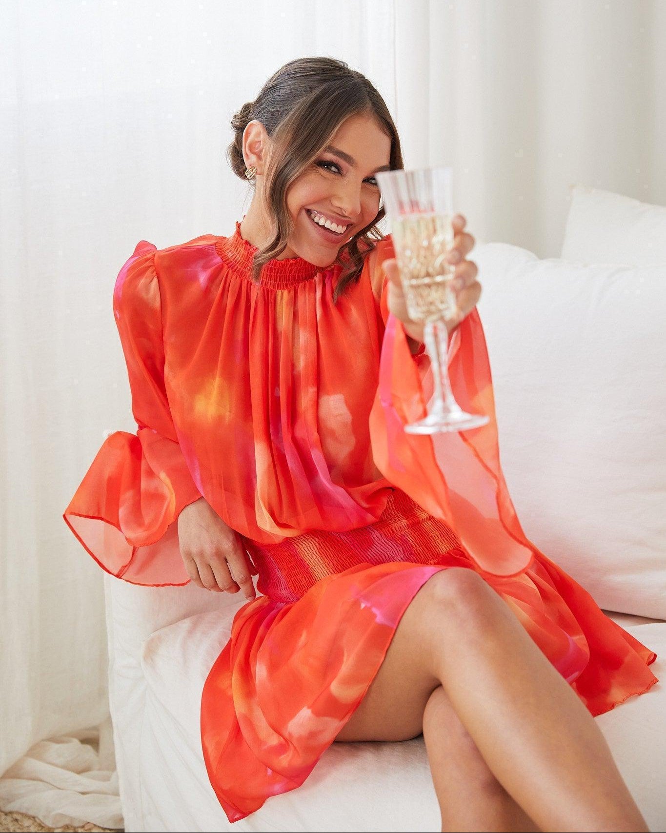 Woman sitting on white couch wearing the Daenerys Orange Print Mini Dress with high neckline and long trumpet sleeves