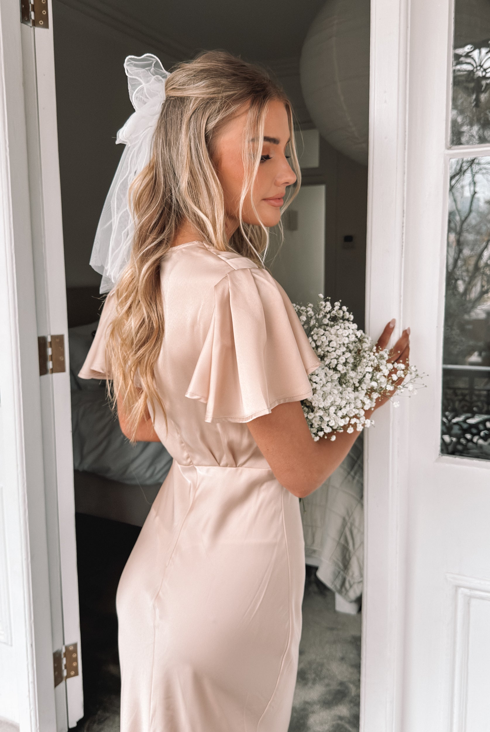 Graceful woman in a beige dress holding a bouquet, suited for wedding celebrations, high-society gatherings, charity dinners, luxury banquets, and elegant affairs, standing in a doorway.