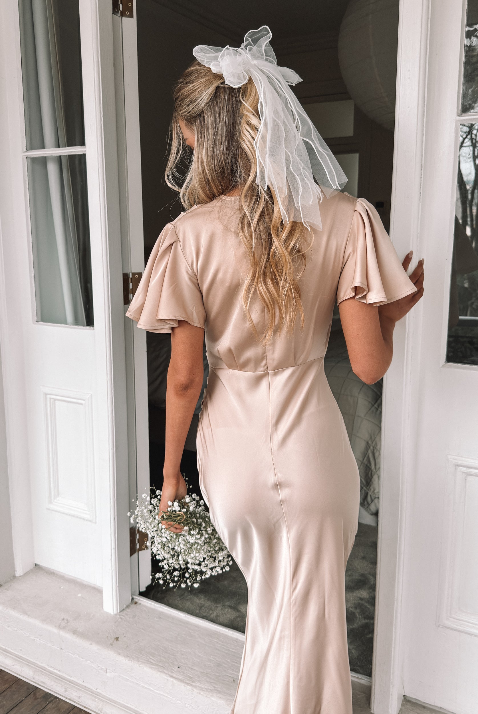Graceful woman in a beige dress holding a bouquet, suited for wedding celebrations, high-society gatherings, charity dinners, luxury banquets, and elegant affairs, standing in a doorway.