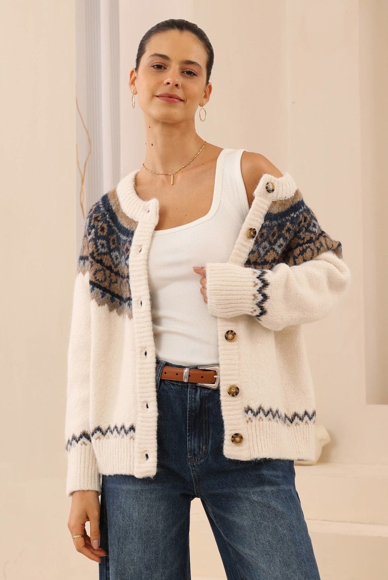 Woman wearing a patterned cardigan over a white top and blue jeans on a neutral background