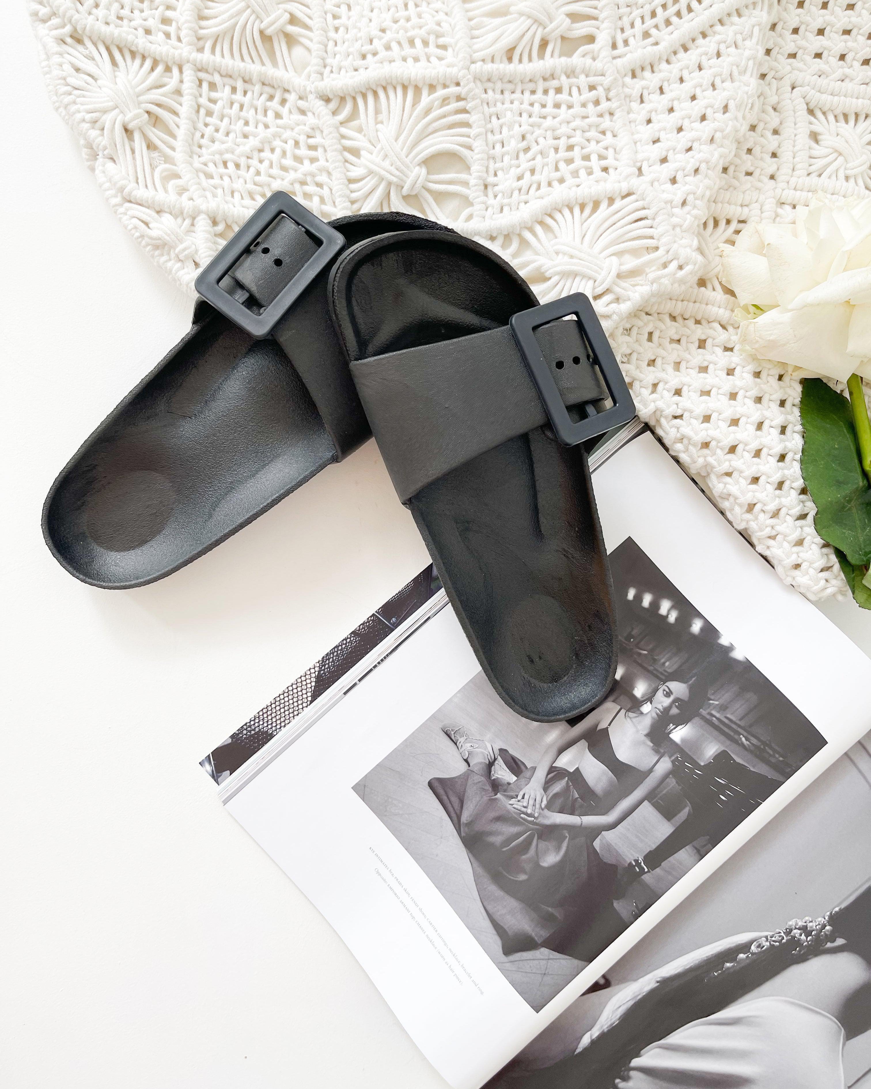 A pair of black summer slides with thick straps, displayed on a flat surface with a knitted texture in the background.