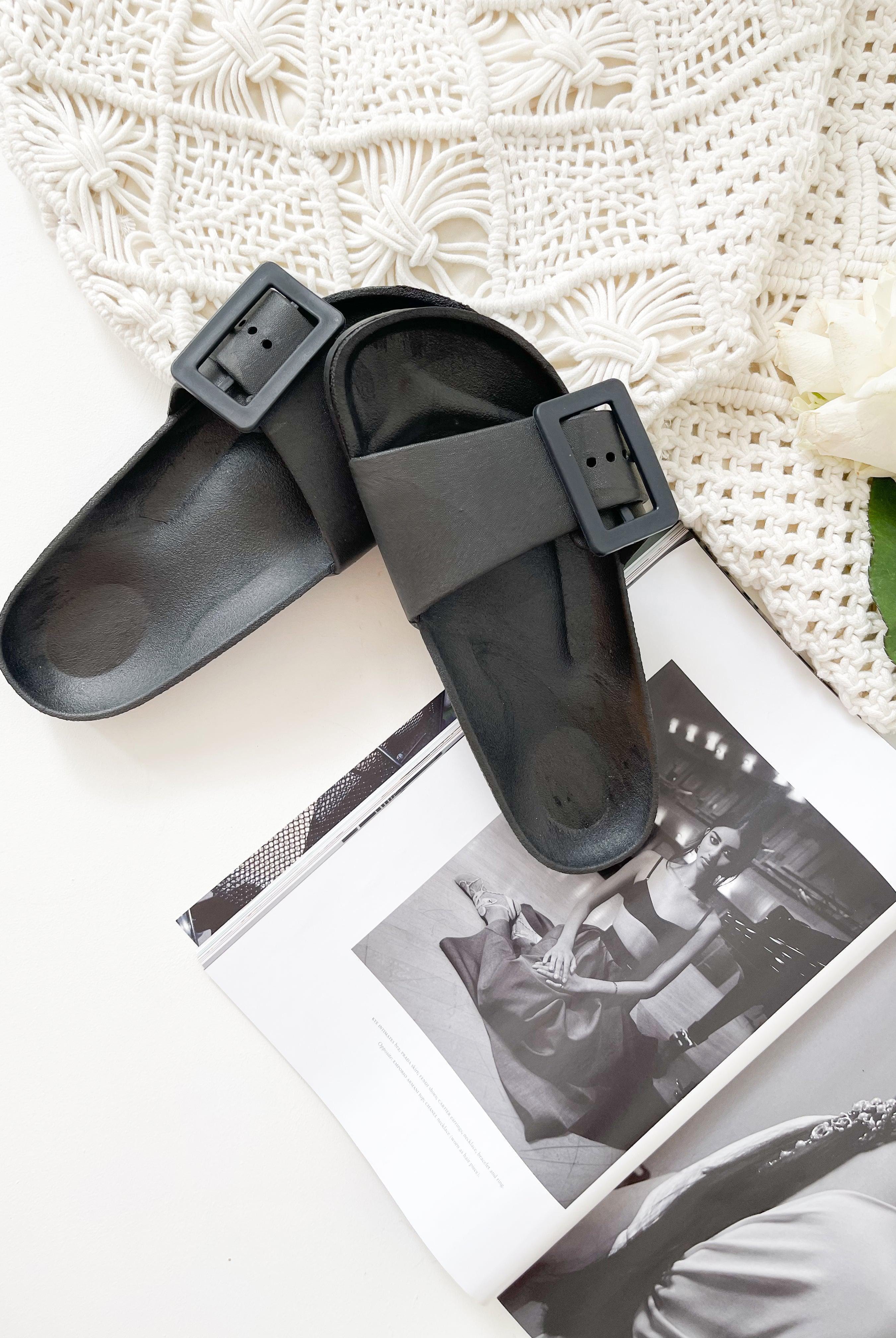A pair of black summer slides with thick straps, displayed on a flat surface with a knitted texture in the background.