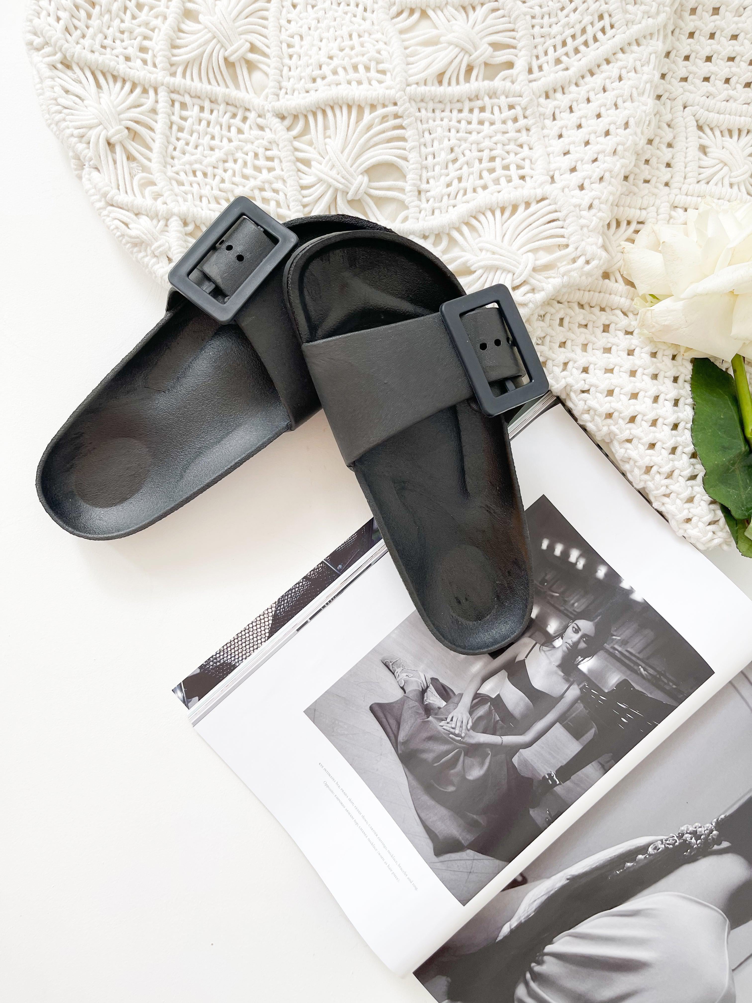 A pair of black summer slides with thick straps, displayed on a flat surface with a knitted texture in the background.