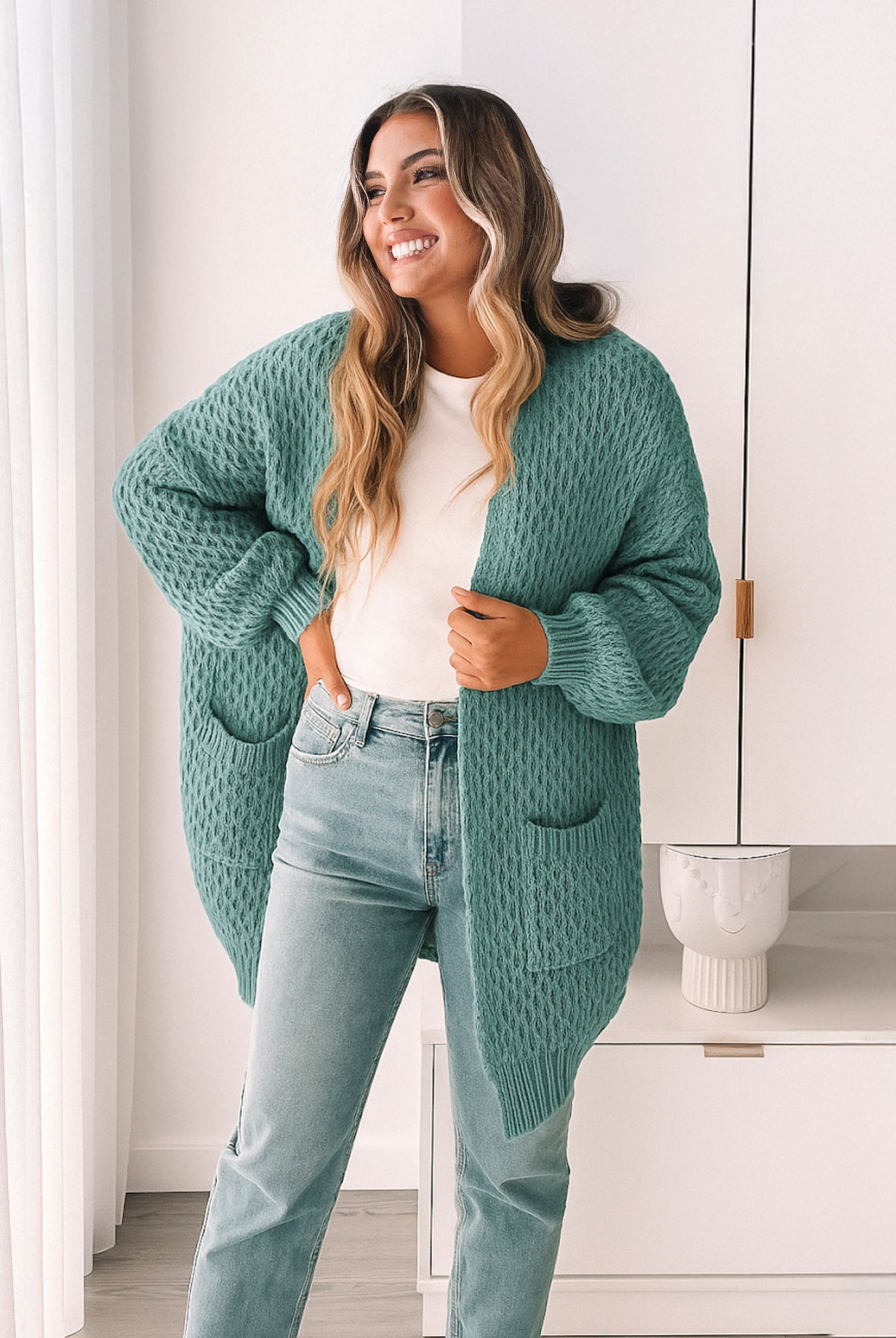Woman wearing a teal cardigan over a white shirt and light blue jeans in a room with white walls and a white cabinet.