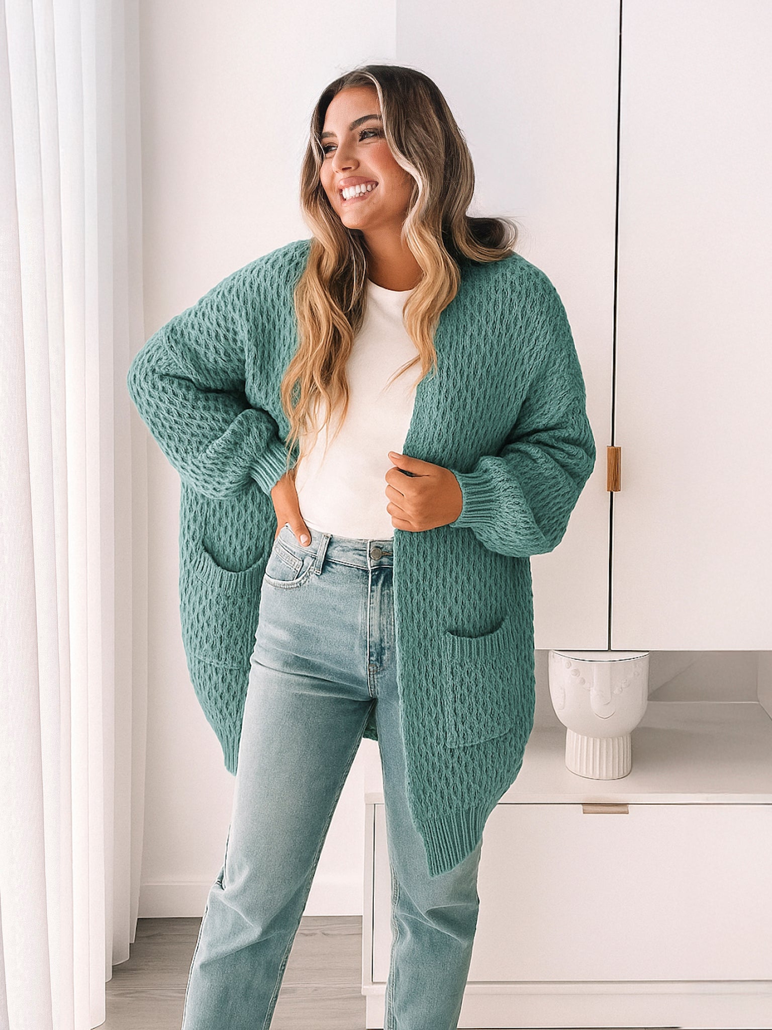 Woman wearing a teal cardigan over a white shirt and light blue jeans in a room with white walls and a white cabinet.