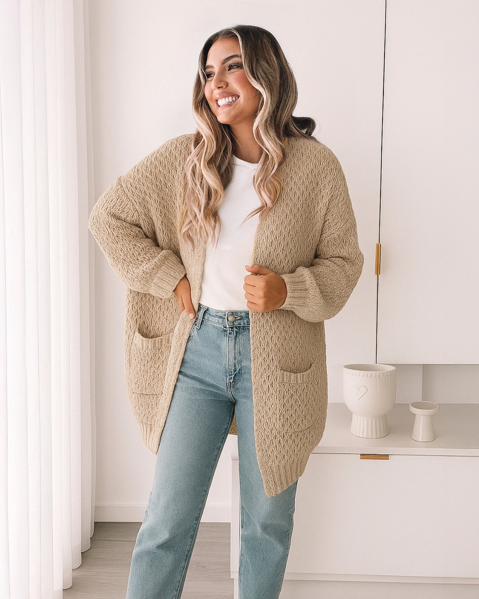 Woman wearing a beige cardigan over a white shirt and light blue jeans in a bright room.