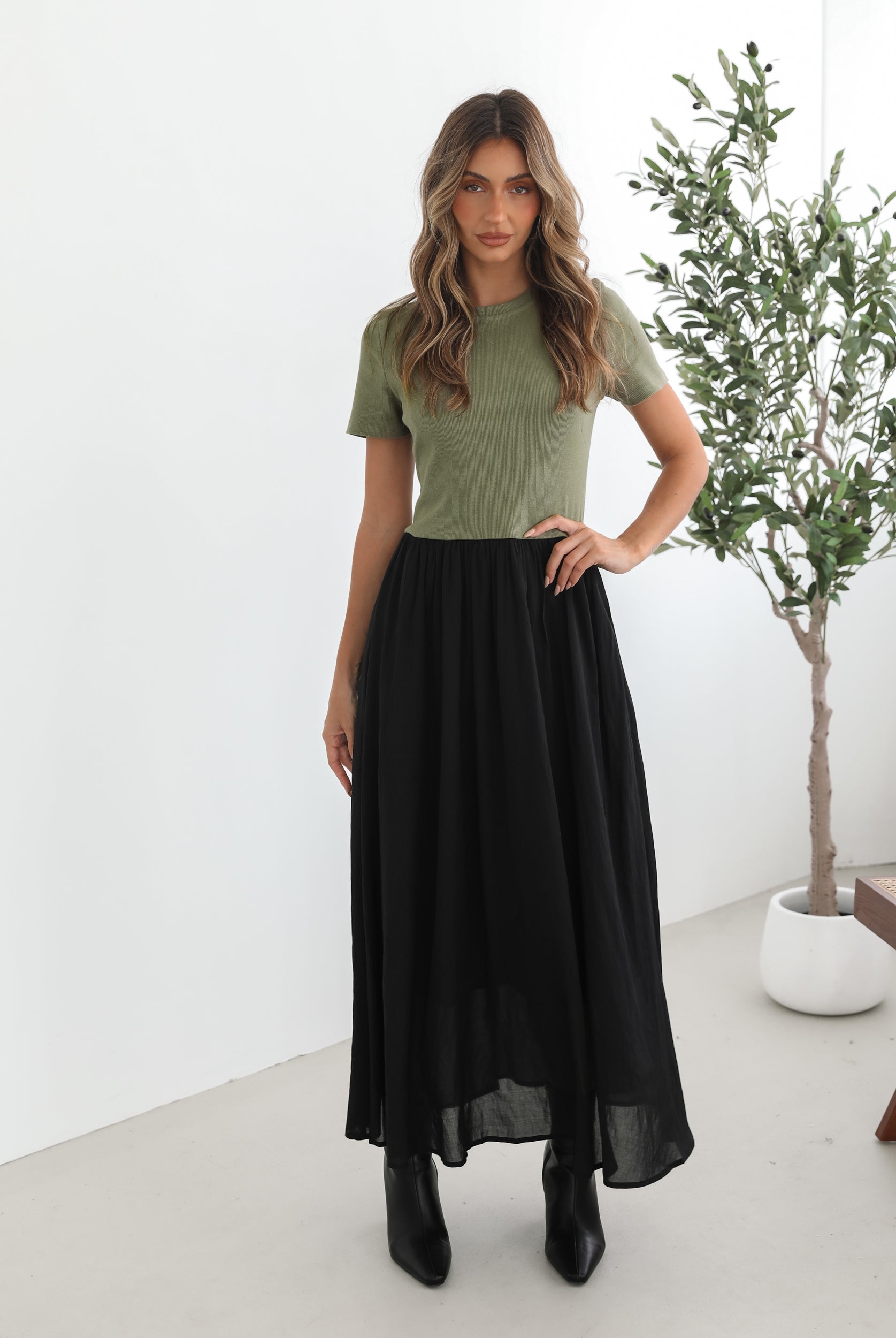 woman wearing a short sleeve round neck dress with green top and black flowy skirt paired with black boots in a white background studio
