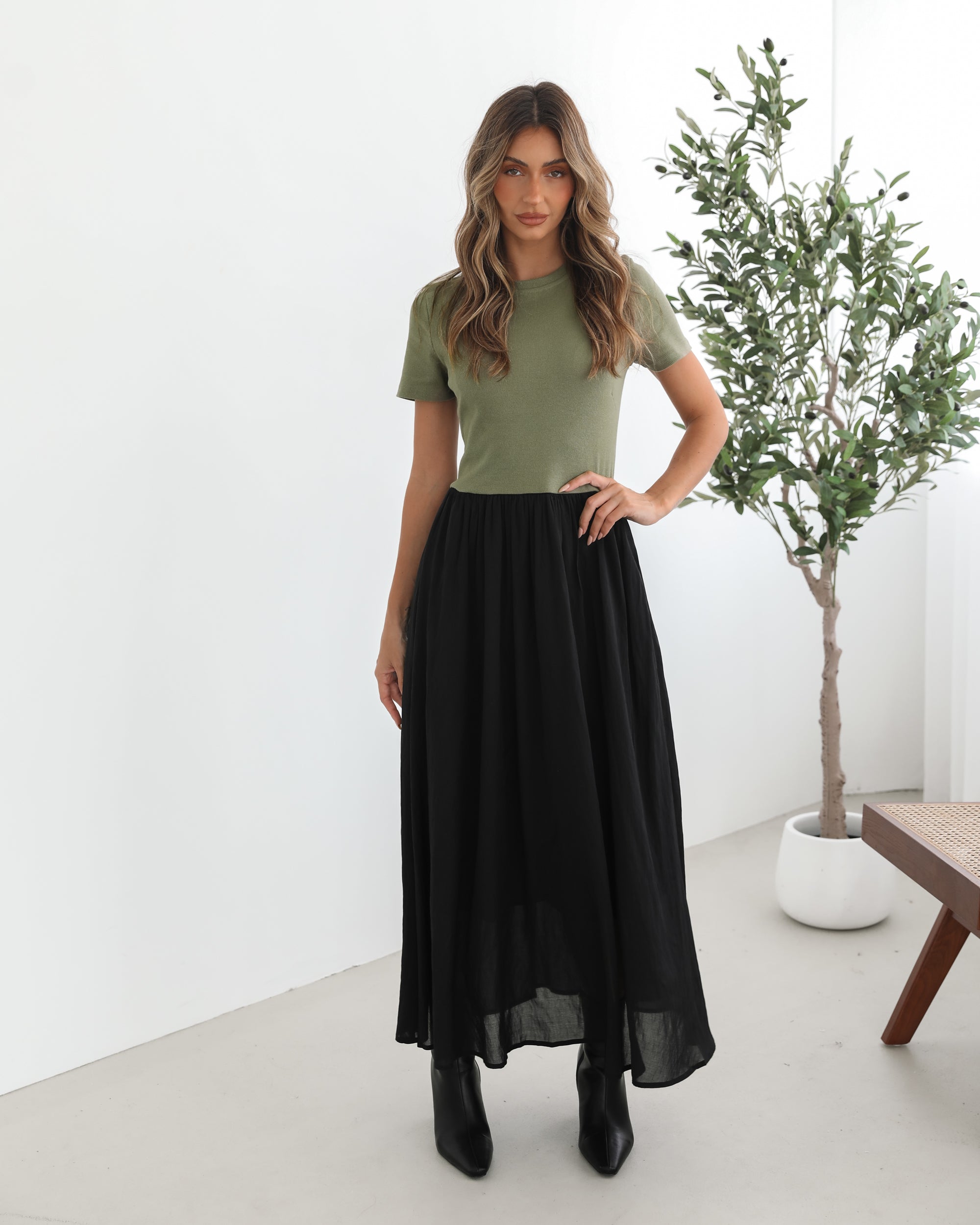 woman wearing a short sleeve round neck dress with green top and black flowy skirt paired with black boots in a white background studio